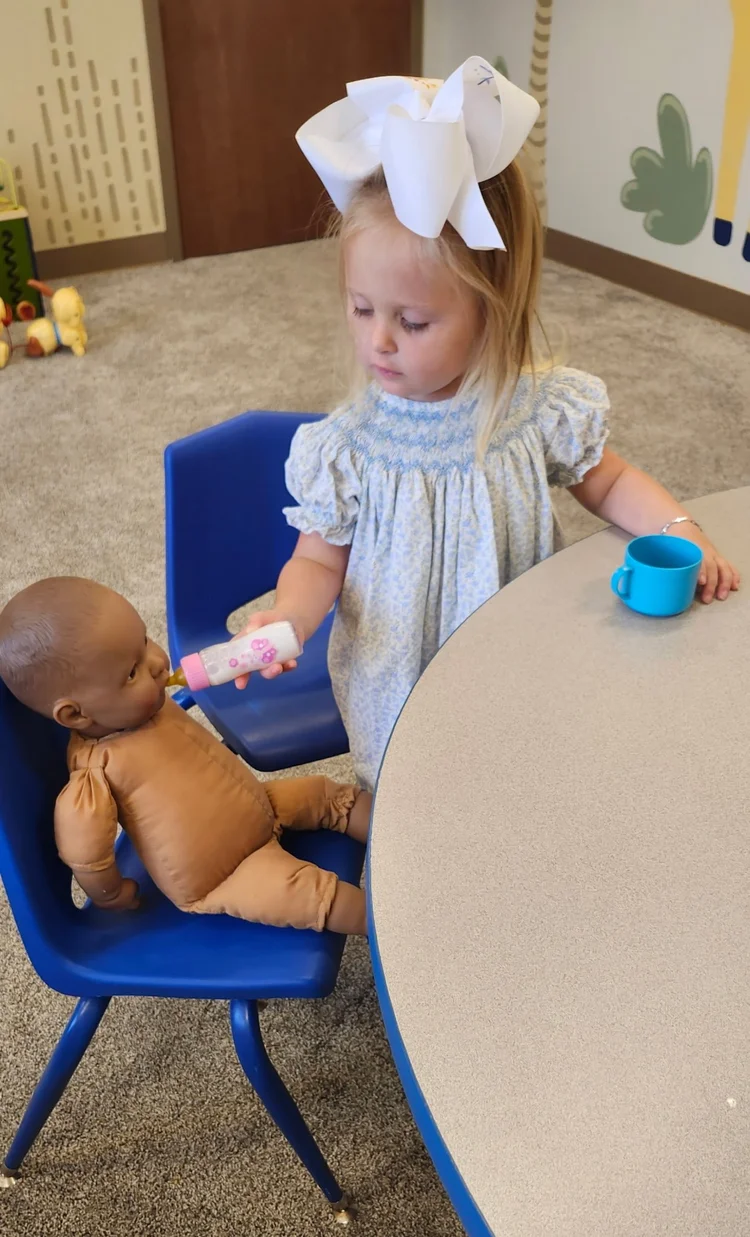 A little girl in a big white bow feeding a baby doll in the preschool room