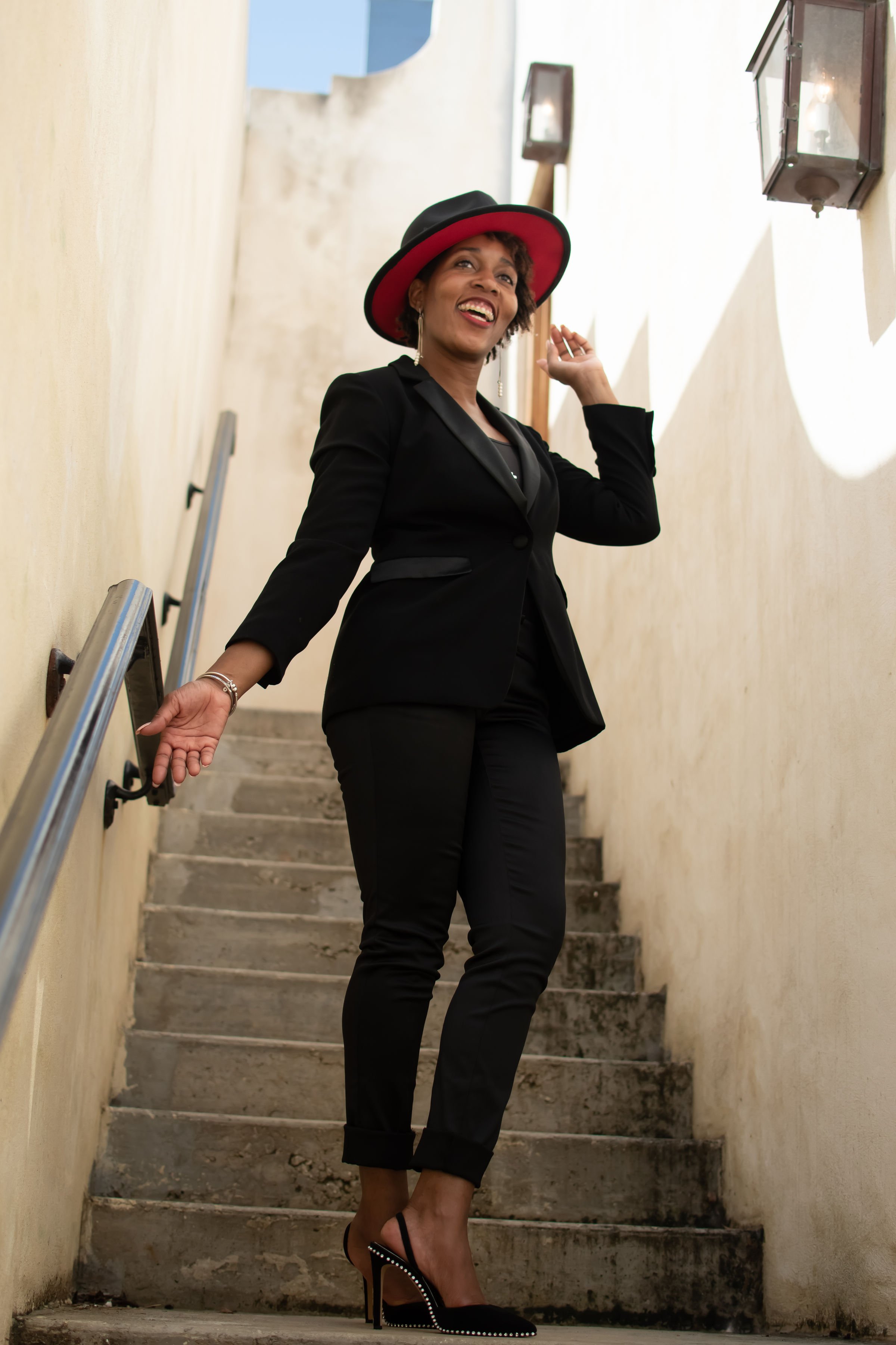 A woman in a black suit, high heels, and a black and red hat walking up a narrow outdoor staircase while smiling and looking to the side.