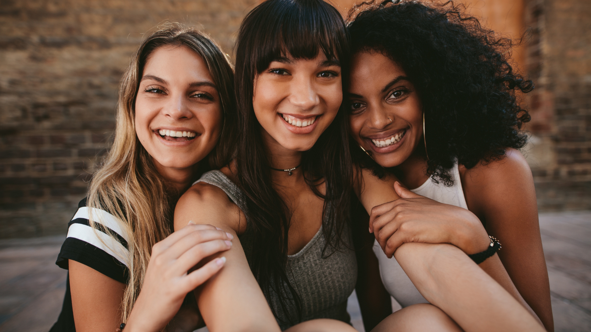 Three women smiling and taking a selfie together indoors, with a brick wall background.