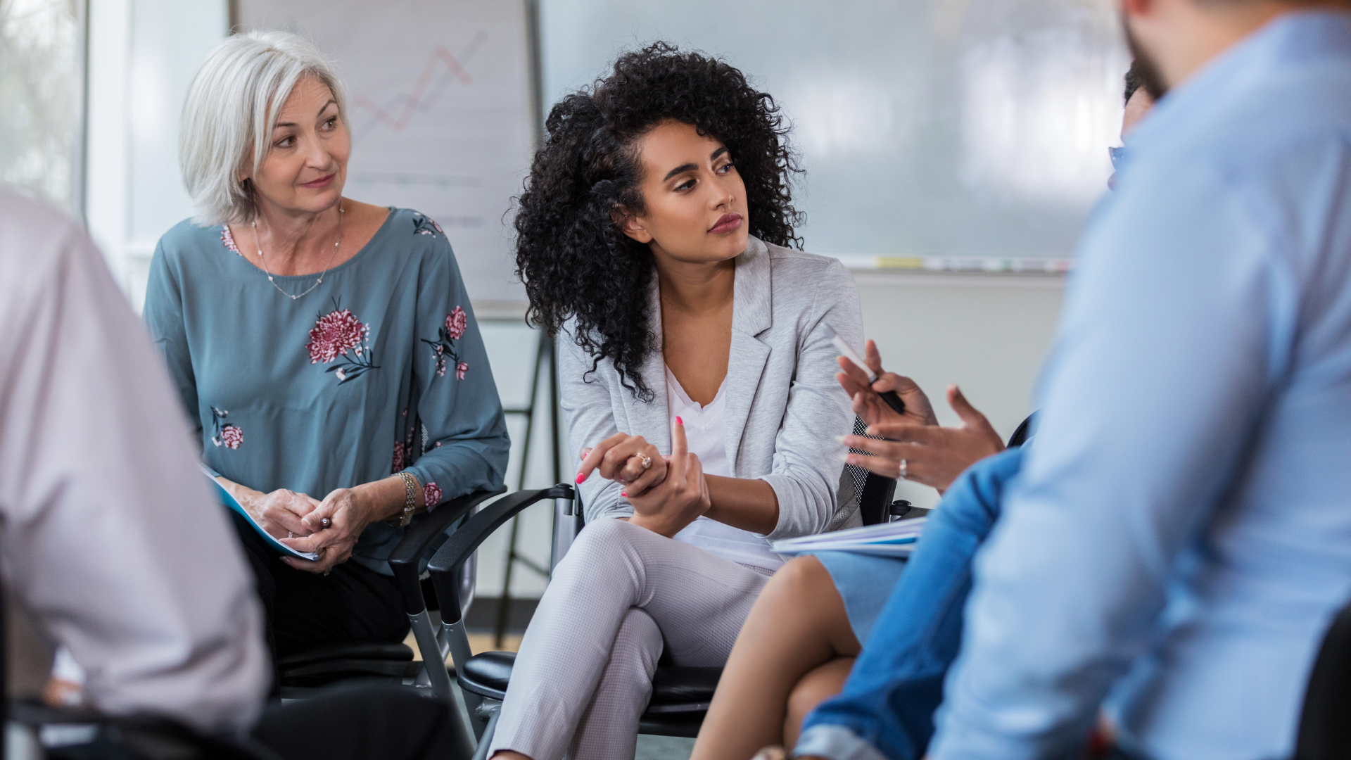A diverse group of business professionals are having a discussion in a conference room, with some holding pens and notebooks.