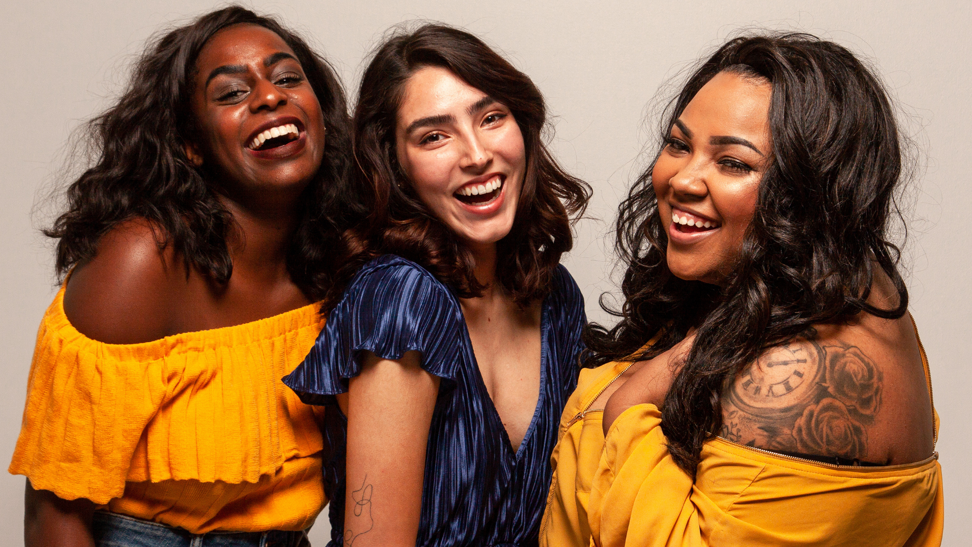 Three women smiling and laughing together, wearing colorful tops, against a plain background.