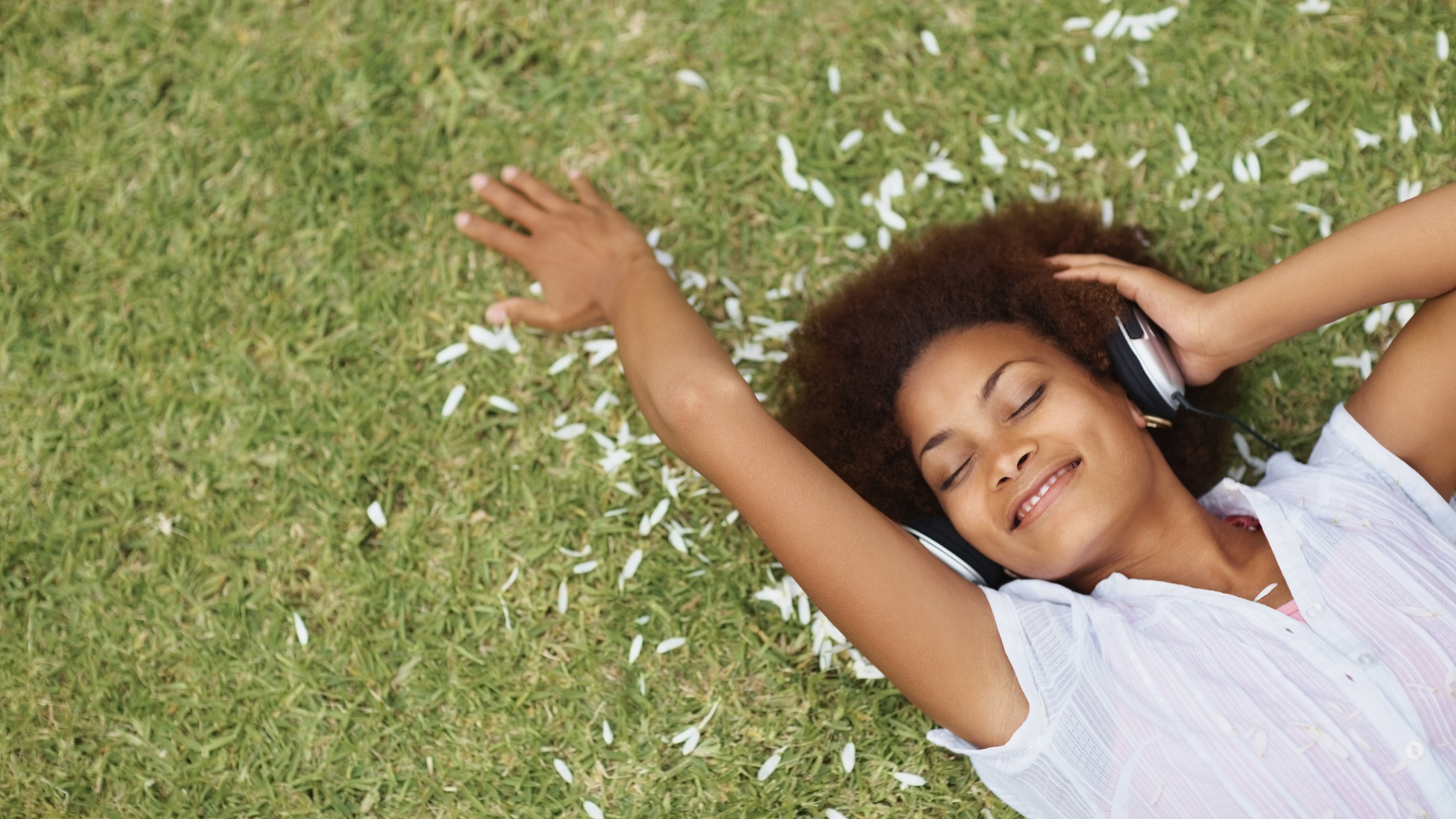 Woman lying on grass, smiling with eyes closed, wearing headphones, surrounded by flower petals.