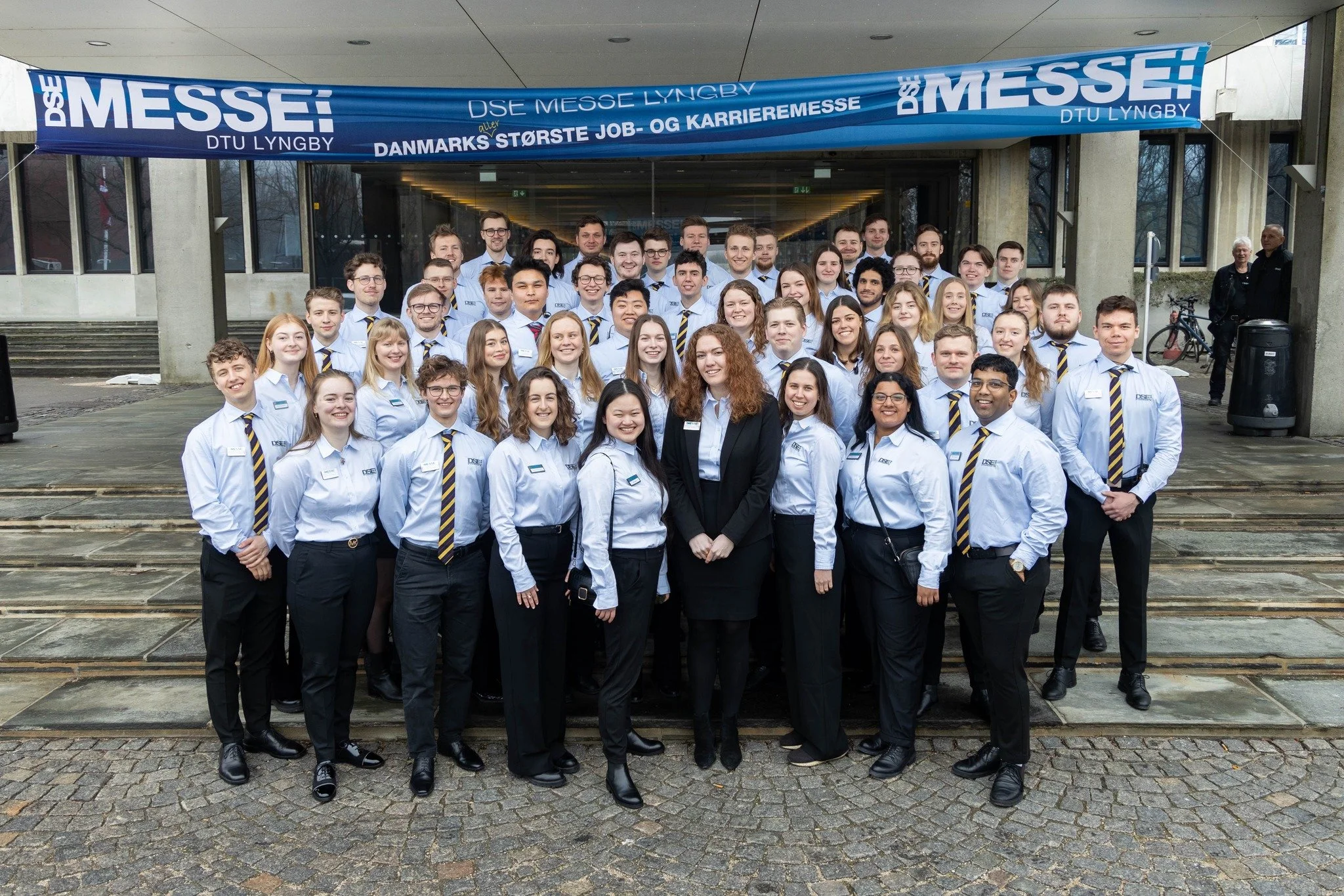 A large group of young professionals, mostly women and men, wearing white shirts and striped ties, standing in front of a building with steps. They are posing for a group photo during an event, with a blue banner overhead that reads "DSE MESSE" and some text in Danish.