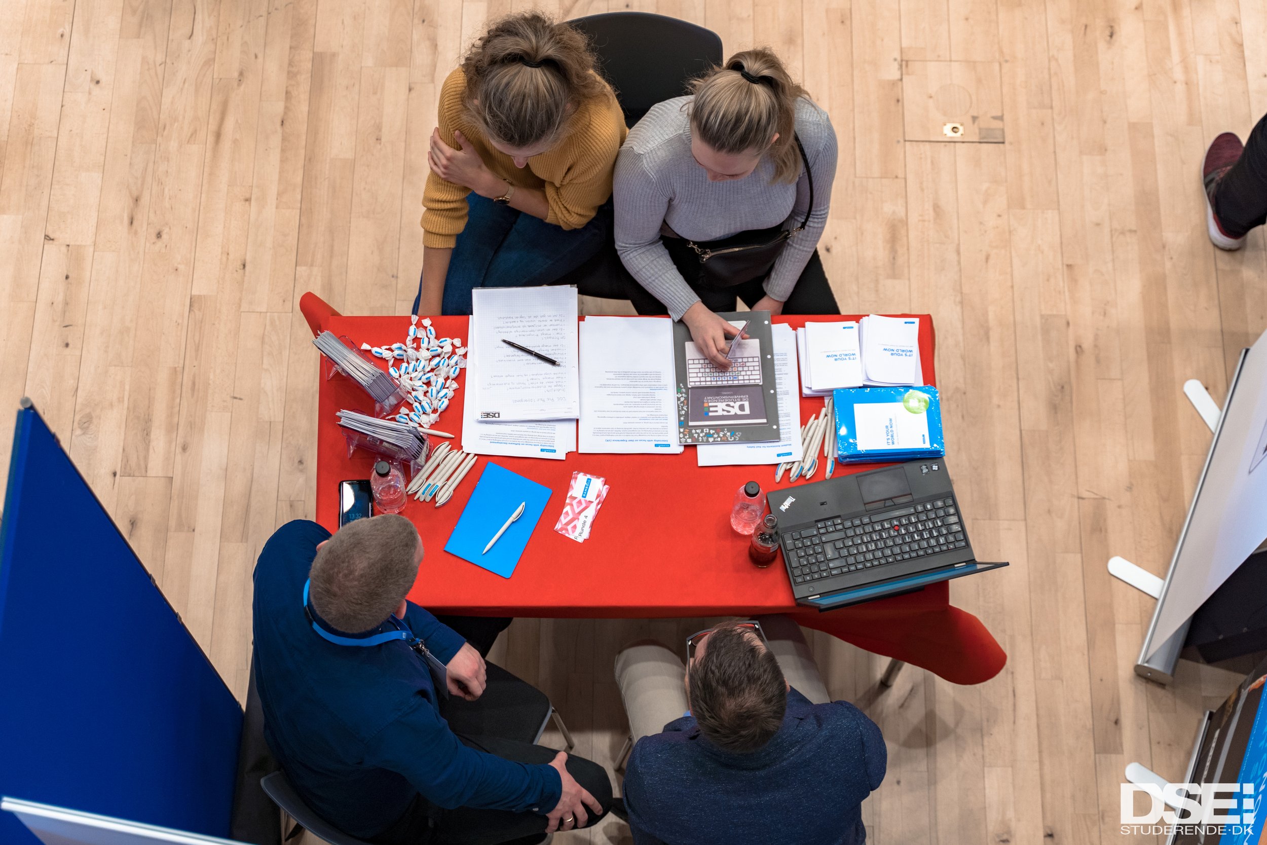 People at a booth with a red table, papers, flyers, pens, and promotional items at an event or conference.