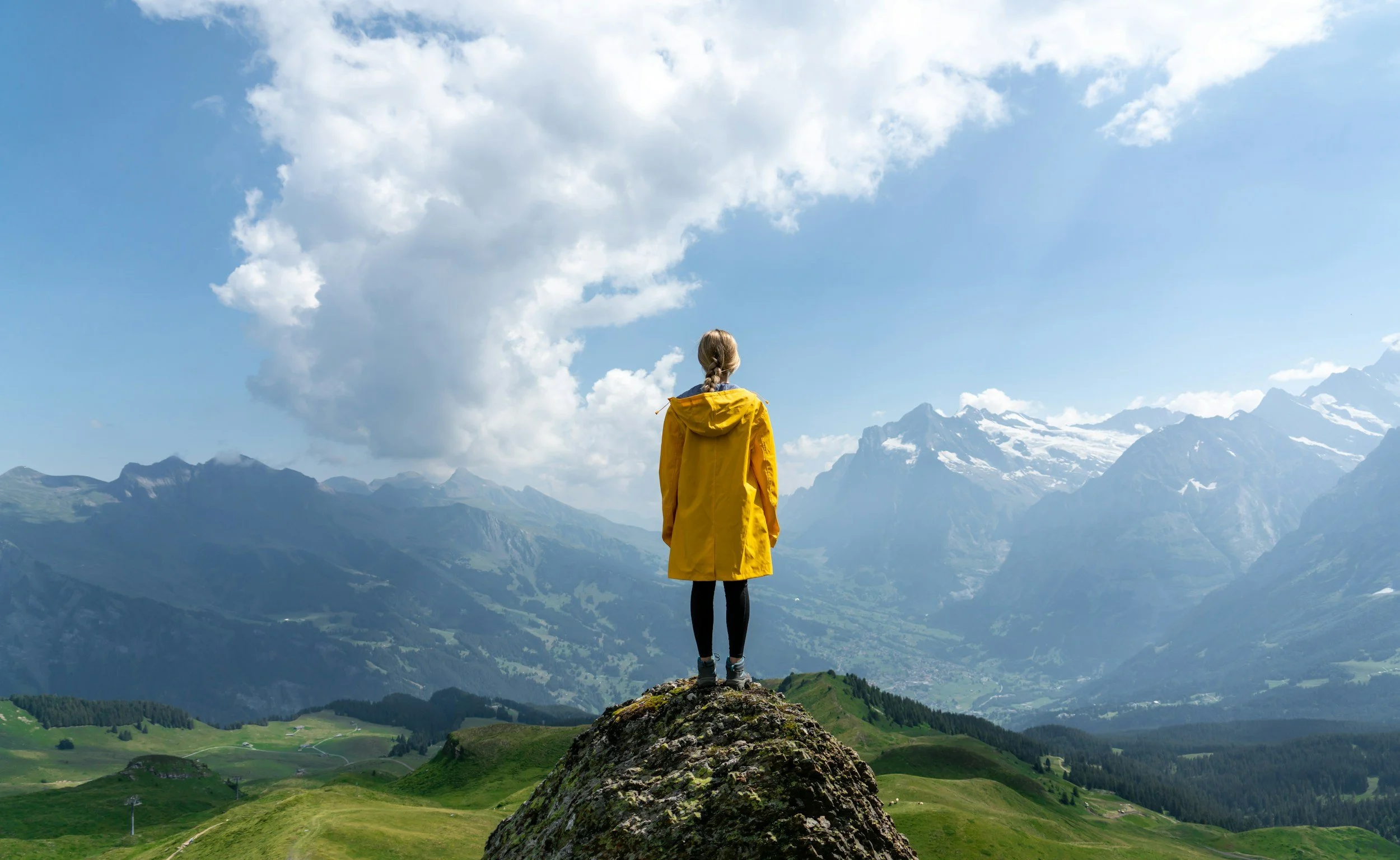 Woman on a mountain top looking at the mountain range in front of her on a sunny day