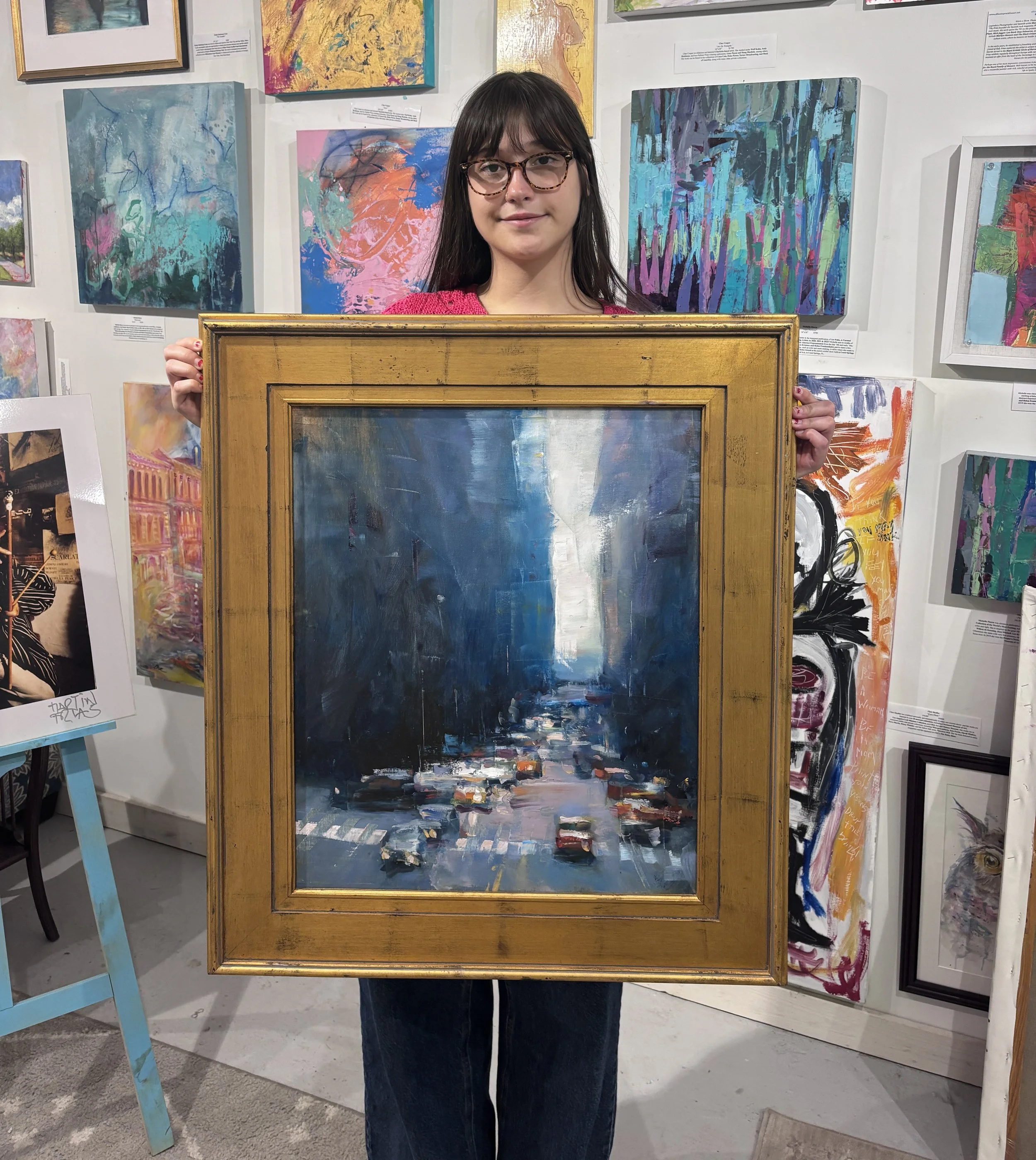 A young woman with dark hair, glasses, and a pink sweater holding a framed abstract painting while standing in an art gallery surrounded by various colorful abstract artworks.