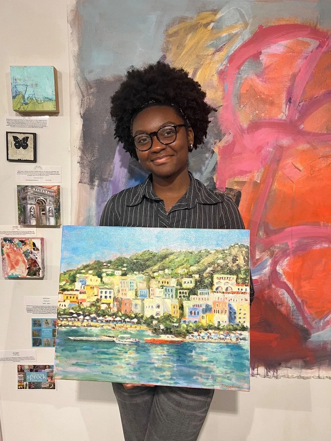 A woman with dark curly hair and glasses holding a colorful painting of a seaside town with hillside buildings and boats in the water. She is standing in front of an abstract mural with pink and orange hues.
