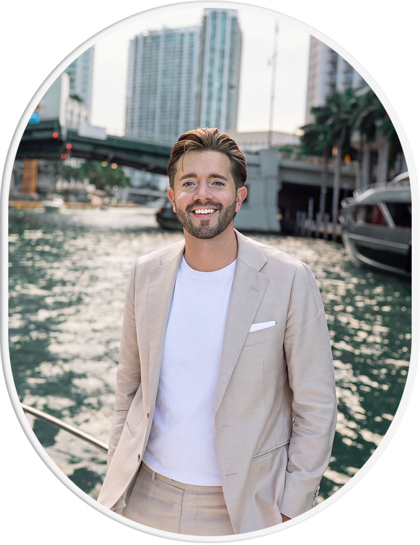 A man with a beard and brown hair smiling while standing by a waterway in an urban area with high-rise buildings and boats in the background.