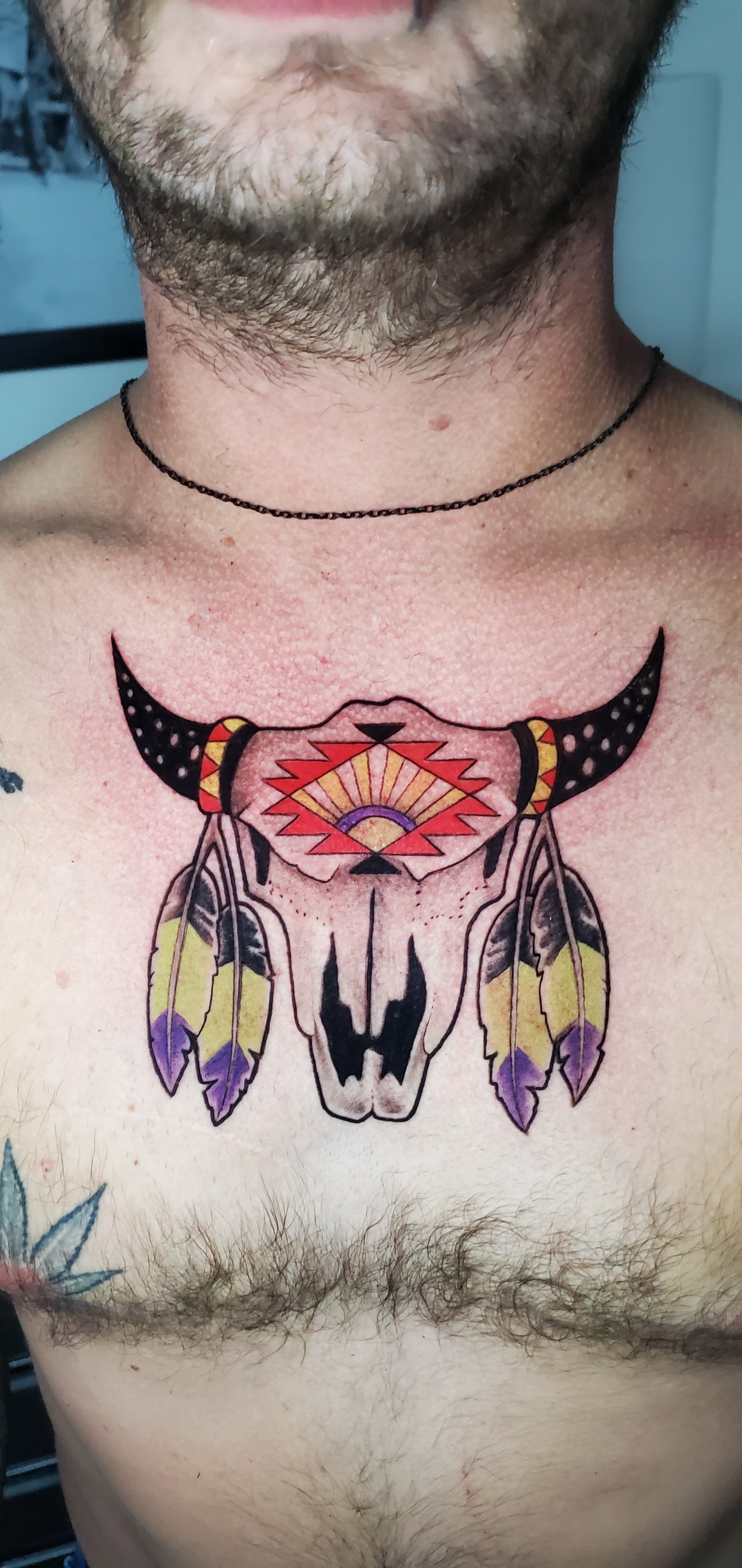 Colorful tattoo of a cow skull with feathers and geometric designs on the chest beneath a necklace and beard.