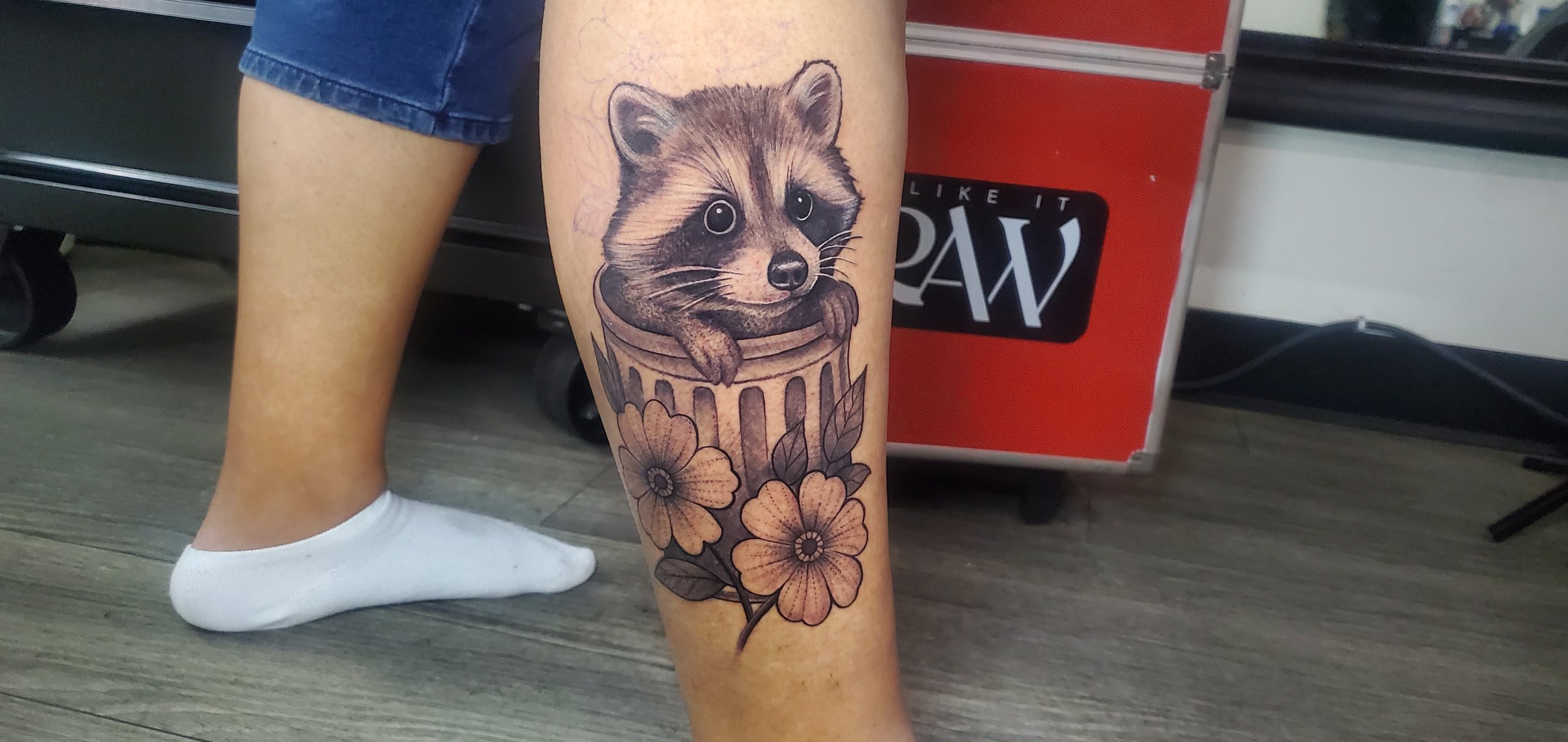 Tattoo of a raccoon peeking out of a trash can, with flowers at the bottom.