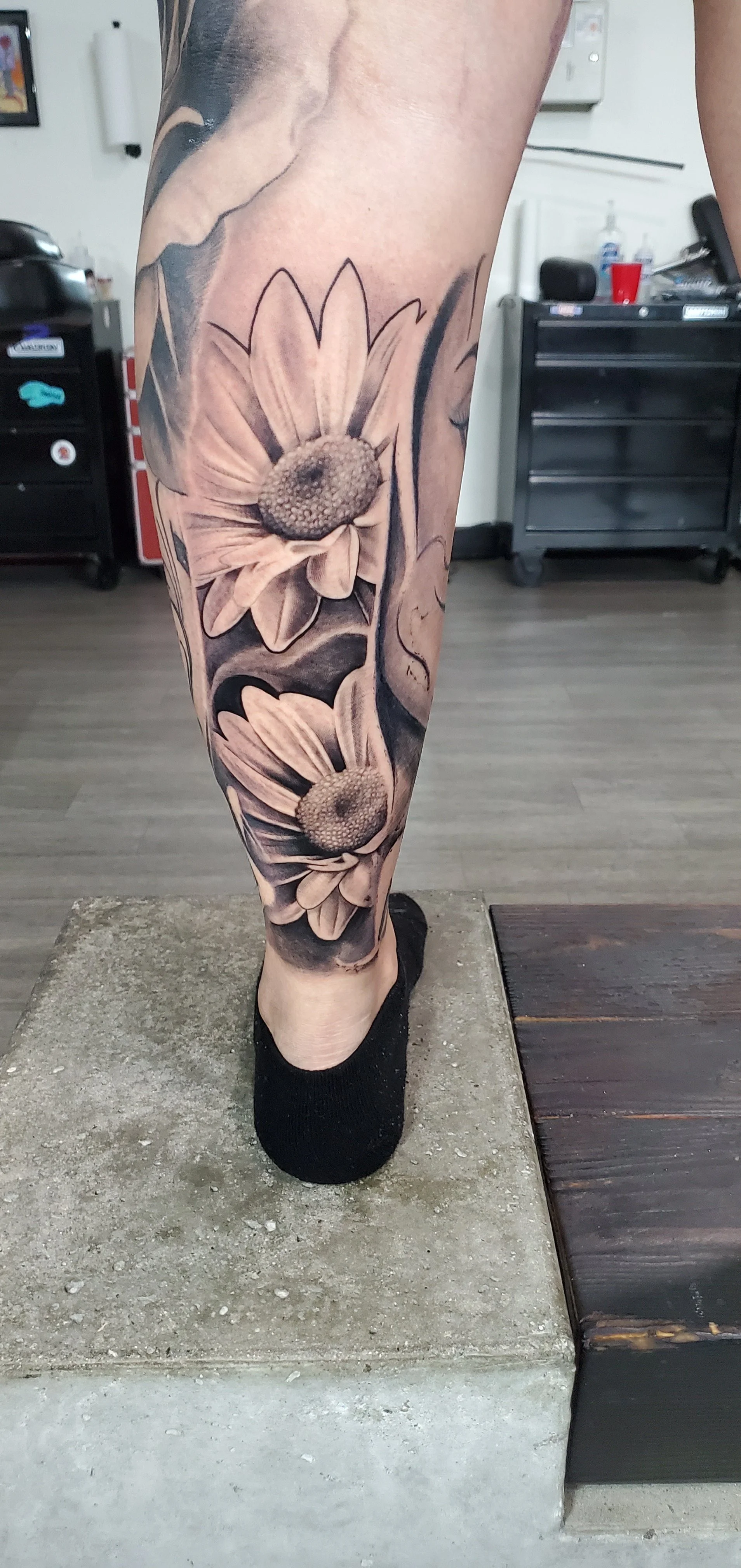 Black and gray floral tattoo of daisies on a person's lower leg.