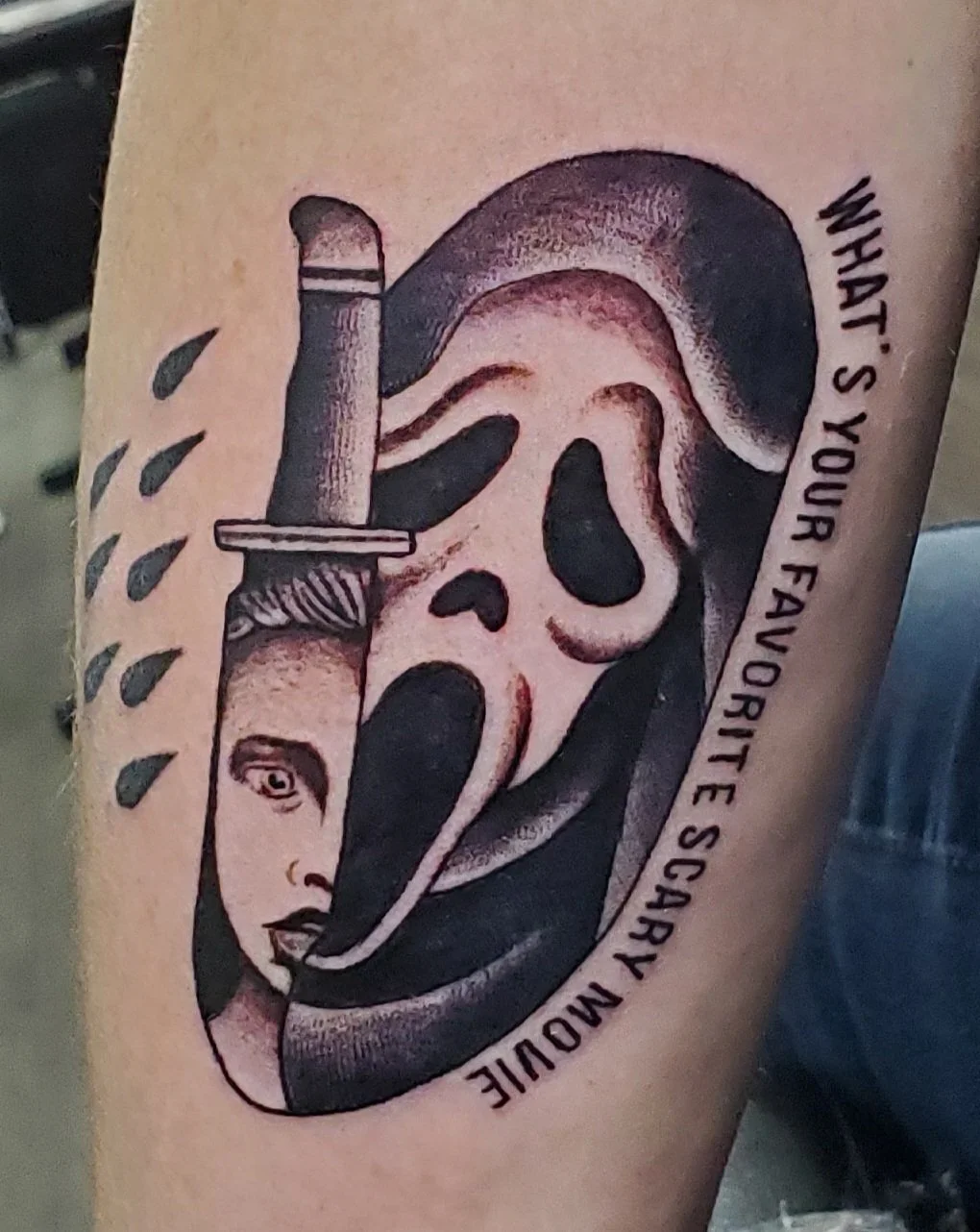 A tattoo of a skull with a tongue sticking out, holding a syringe with a small, detailed portrait of a woman's face inside the tongue. The tattoo includes the text "WHAT'S YOUR FAVORITE SCARY MOVIE" curved around the skull.