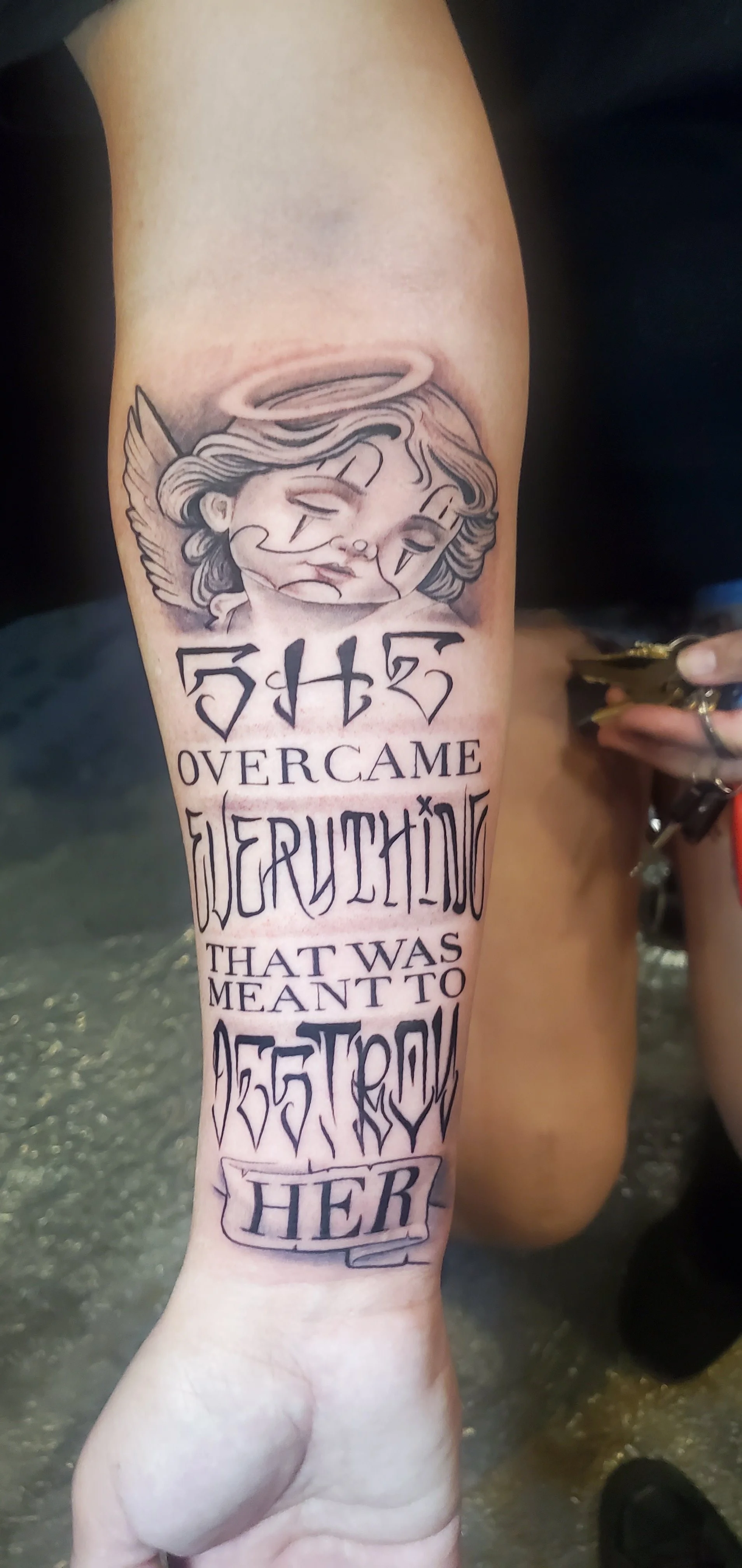 Tattoo of a crying angel with a halo on the upper arm, with text that reads, "Overcame everything that was meant to destroy her."