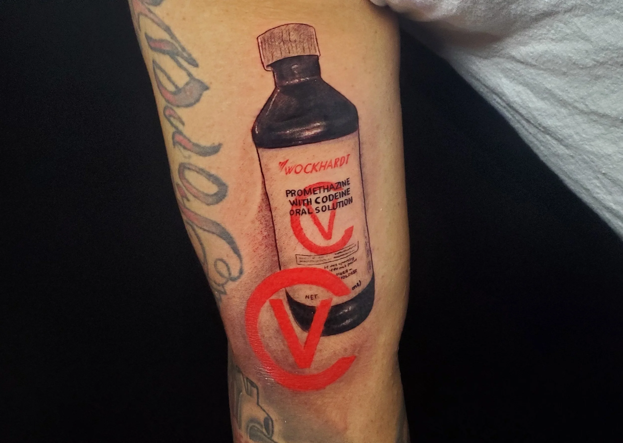 Tattoo of a black medicine bottle with a wooden cap, labeled 'Wockhardt' with additional text 'Promethazine with Codeine Oral Solution' and a red stylized heart design below.