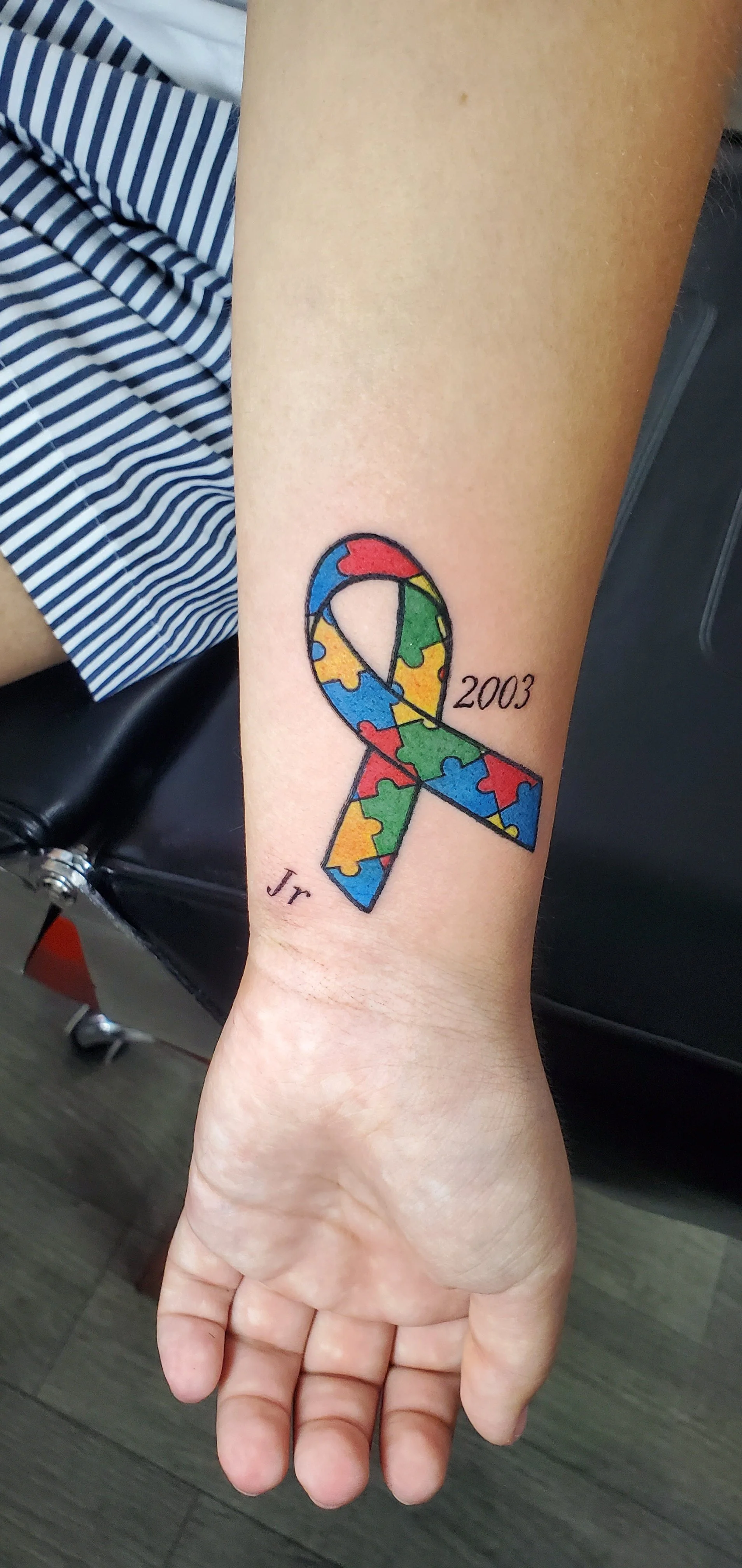 Tattoo of a puzzle piece ribbon with a red, blue, green, and yellow color pattern. The ribbon has the year 2003 and initials Jr. beneath it.