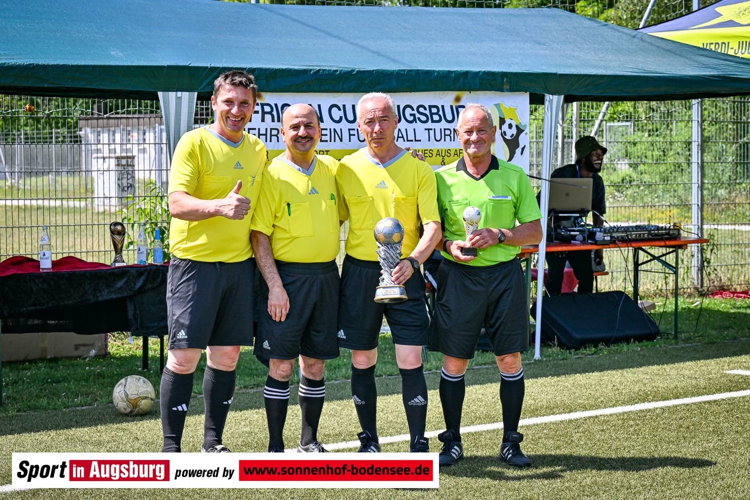 Vier Männer in gelben und grünen Fußballschuhen stehen auf einem Fußballfeld unter einem grünen Zelt. Zwei von ihnen halten Pokale. Hinter ihnen steht eine Tafel mit einem Fußball-Symbol und Text, im Hintergrund sind Zaun und Bäume. Einer der Männer tröstet einen weiteren. Die Szene ist bei einer Fußballveranstaltung in Augsburg.