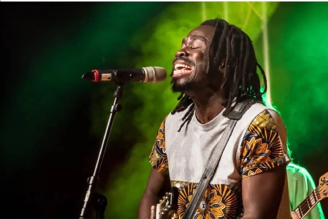 Man singt und spielt Gitarre auf der Bühne, trägt ein farbenfrohes Hemd mit afrikanischen Mustern, bei grüner Bühnenbeleuchtung.