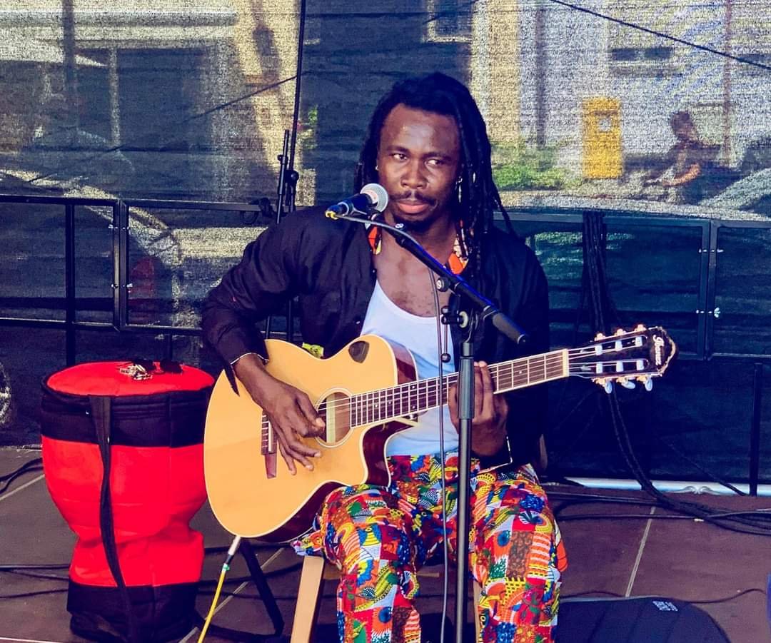 Ein Musiker mit dunkler Haut, Afro-Haar und Dreadlocks sitzt mit einer Gitarre auf einem Stuhl, singt in ein Mikrofon und spielt Musik. Er trägt bunte Hosen mit afrikanischen Mustern und eine schwarze Jacke.