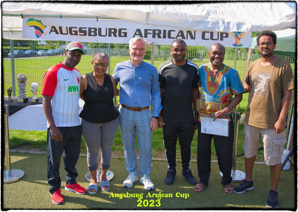 Sechs Menschen stehen unter einem Zelt mit dem Banner "Augsburg African Cup 2023". Im Hintergrund sind Trophäen und eine Grastestfläche sichtbar.