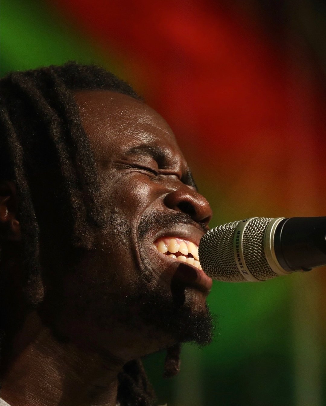 Mann singt in ein Mikrofon, nahe am Gesicht, mit geschlossenen Augen, vor einem bunten, unscharfen Hintergrund.