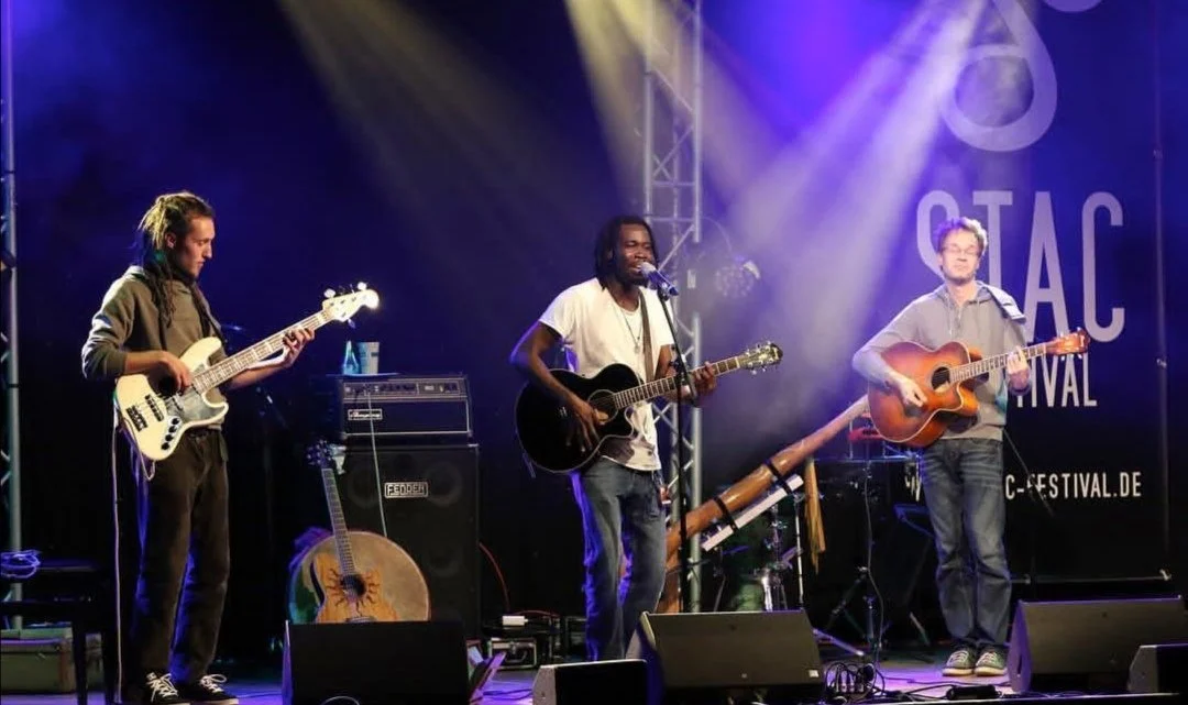 Drei Musiker auf einer Bühne beim Auftritt, spielen Gitarre und Bass, im Hintergrund ein Banner mit der Aufschrift "STAC FESTIVAL"