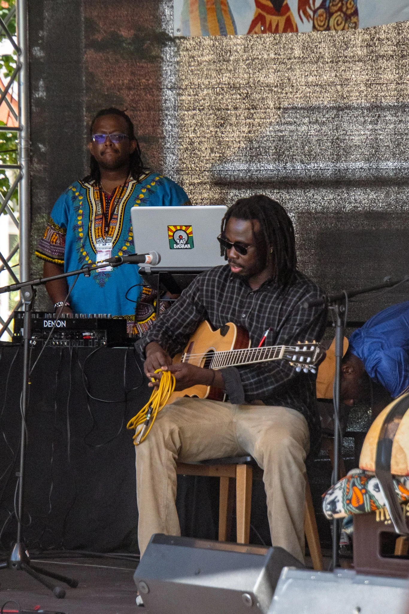 Musiker spielt Gitarre auf einer Bühne bei einem Musikfestival, im Hintergrund ein DJ mit Laptop, beide tragen lässige Kleidung und Sonnenbrillen.