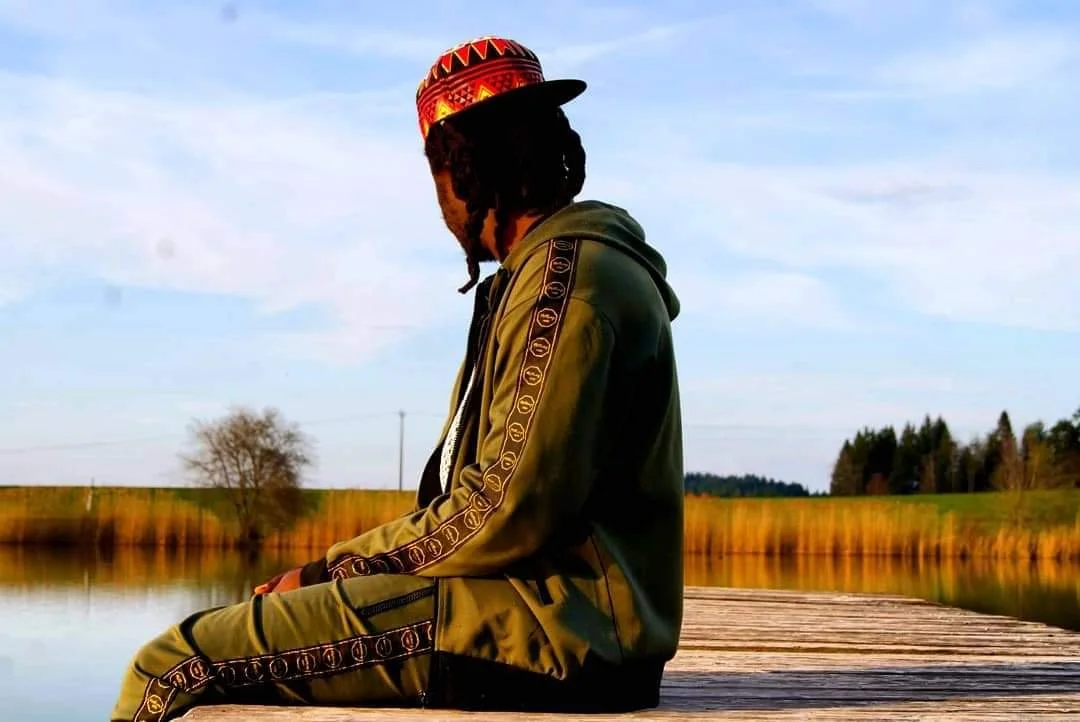 Eine Person sitzt an einem Wasser, trägt einen bunten Hut und eine grüne Jacke, im Hintergrund sind Bäume und ein offener Himmel.