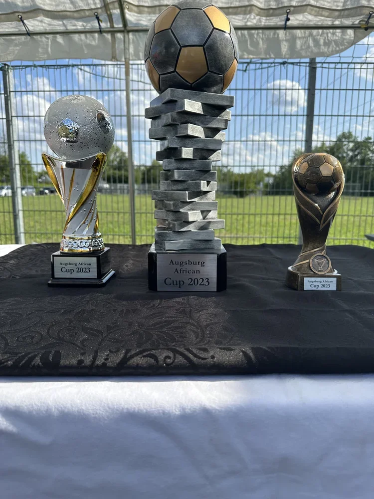 Drei Pokale für die Augsburger African Cup 2023, mit einer großen Trophäe in der Mitte, flankiert von zwei kleineren Pokalen, auf einem schwarzen Tuch.