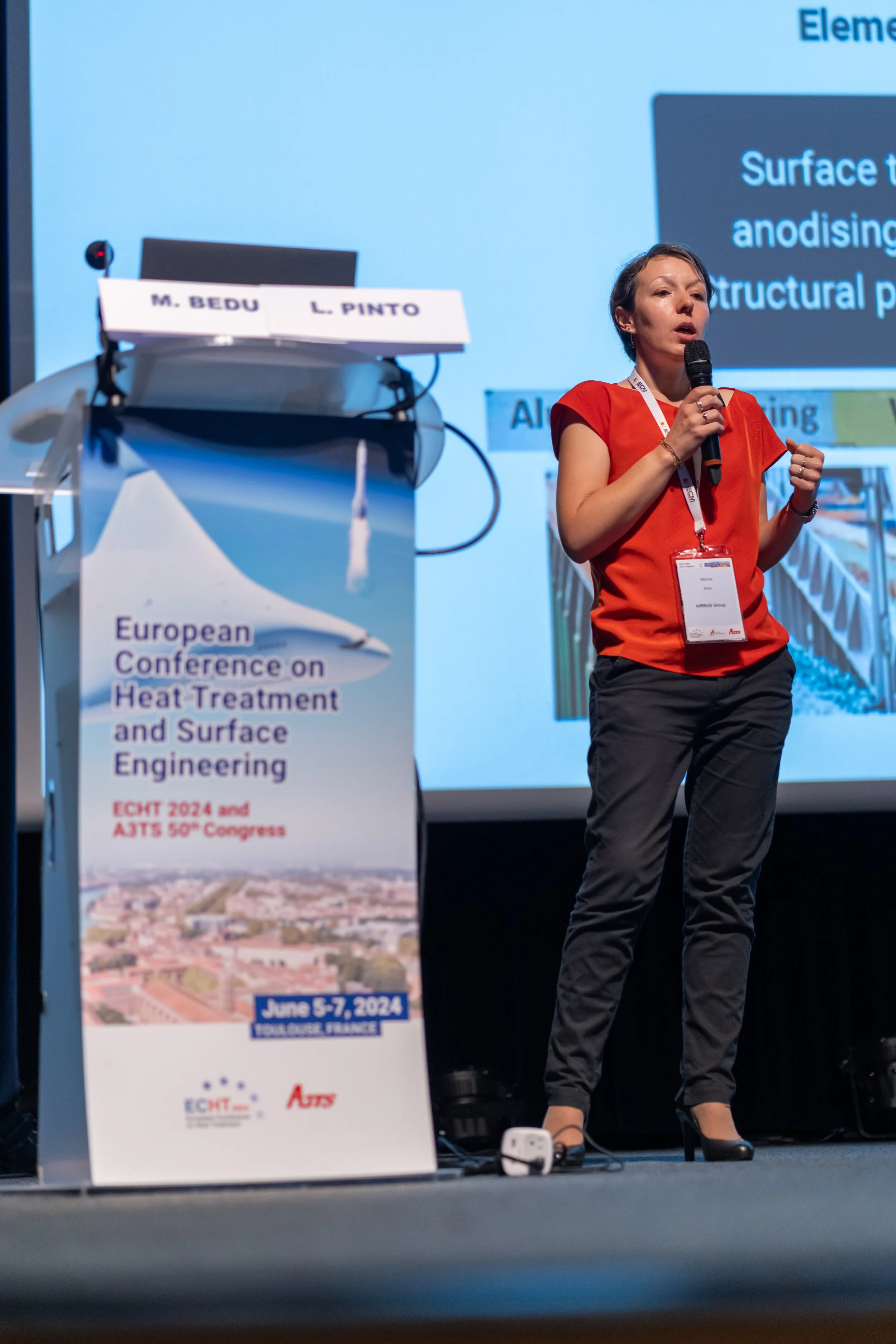 Une femme en chemise rouge tient un micro et parle à une conférence, avec un podium à côté affichant une affiche pour la "Conférence Européenne sur le Traitement Thermique et l’Ingénierie des Surfaces" à Toulouse en 2024.