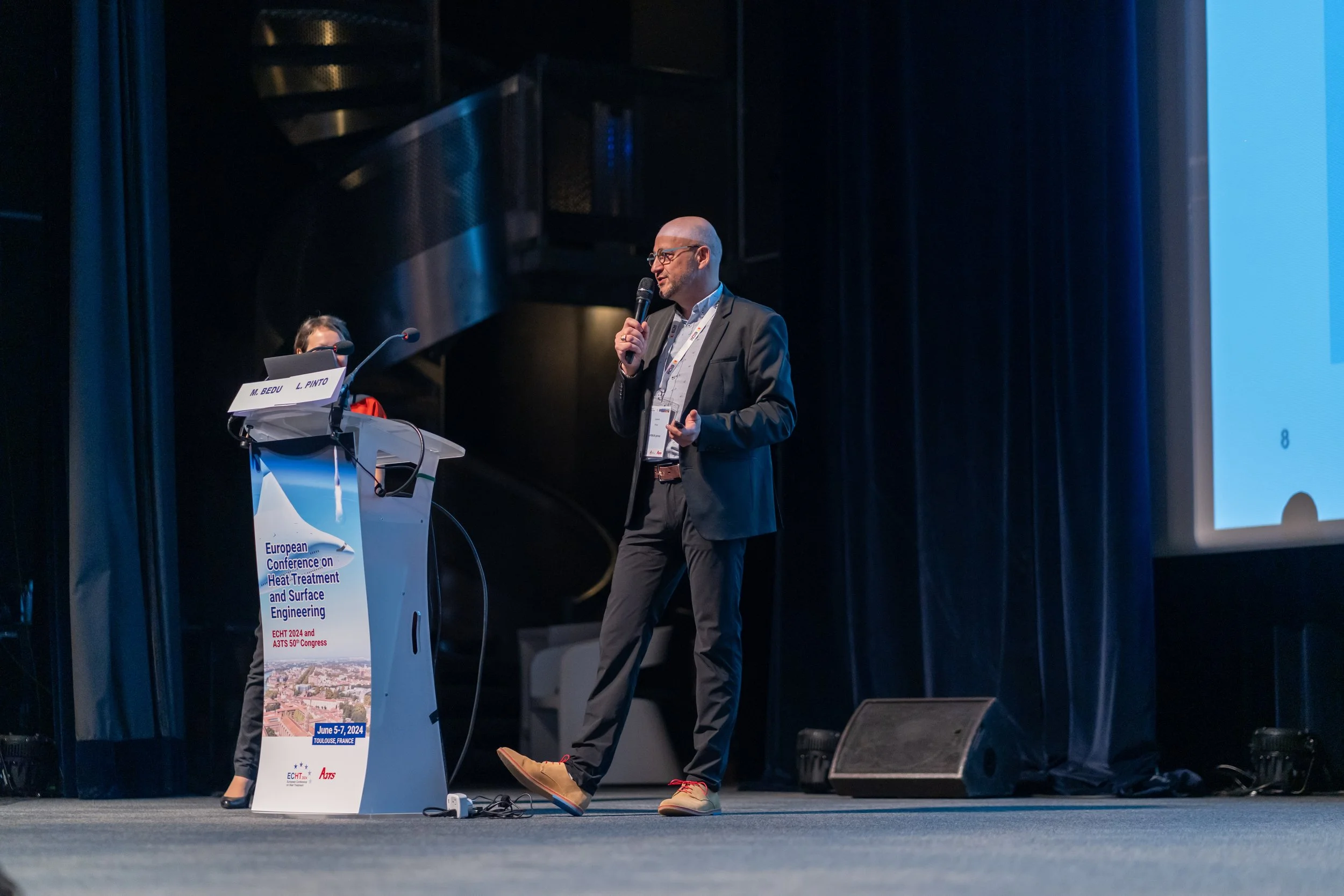 Un homme en costume fait une présentation sur scène avec un micro, devant un pupitre avec l'affiche d'une conférence européenne sur le traitement thermique et l'ingénierie de surface. Un écran de présentation est visible à droite.