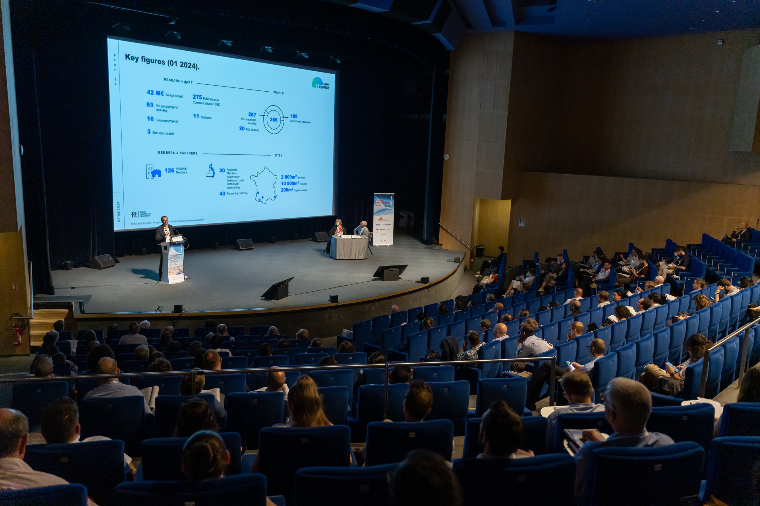 Conférence en salle avec présentation sur grand écran et public assis, intervenants sur scène.