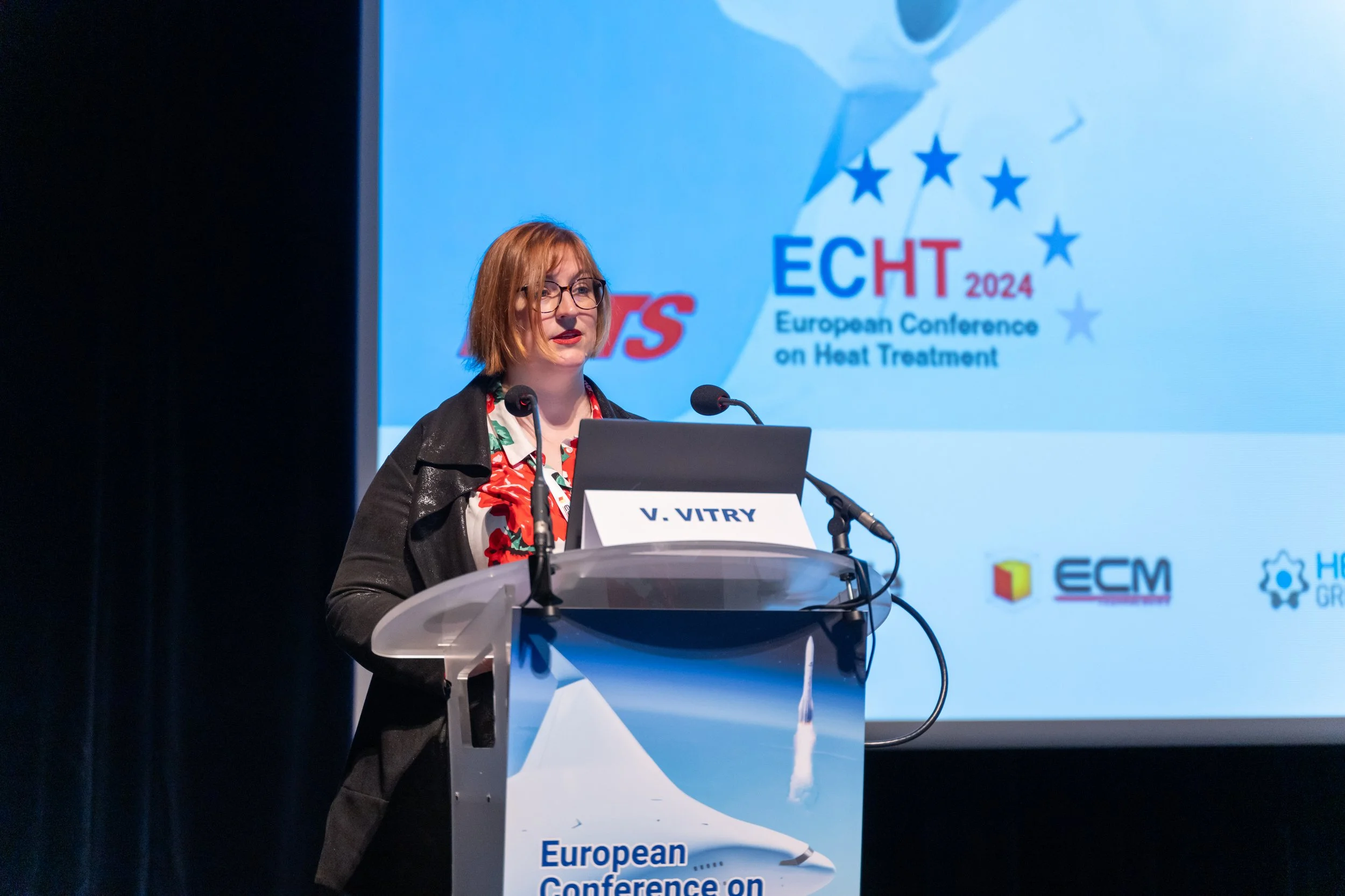 Une personne présente à une conférence européenne sur le traitement thermique, debout à un podium avec des micros et un écran d'affichage à l'arrière-plan.