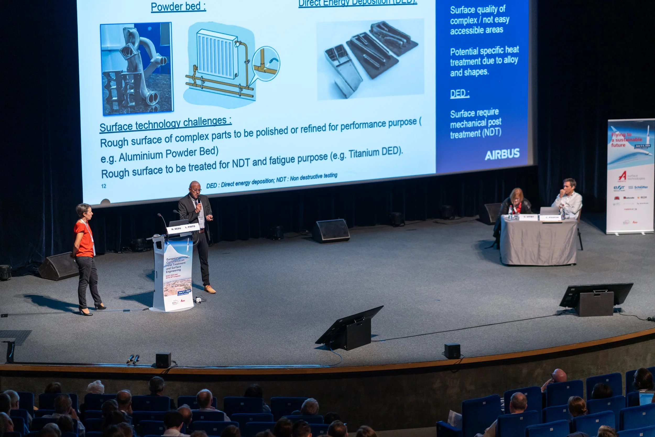 Conférence Airbus avec orateur et diapositives présentant des technologies de surface pour impression 3D. Deux personnes assises à une table, public assis devant.