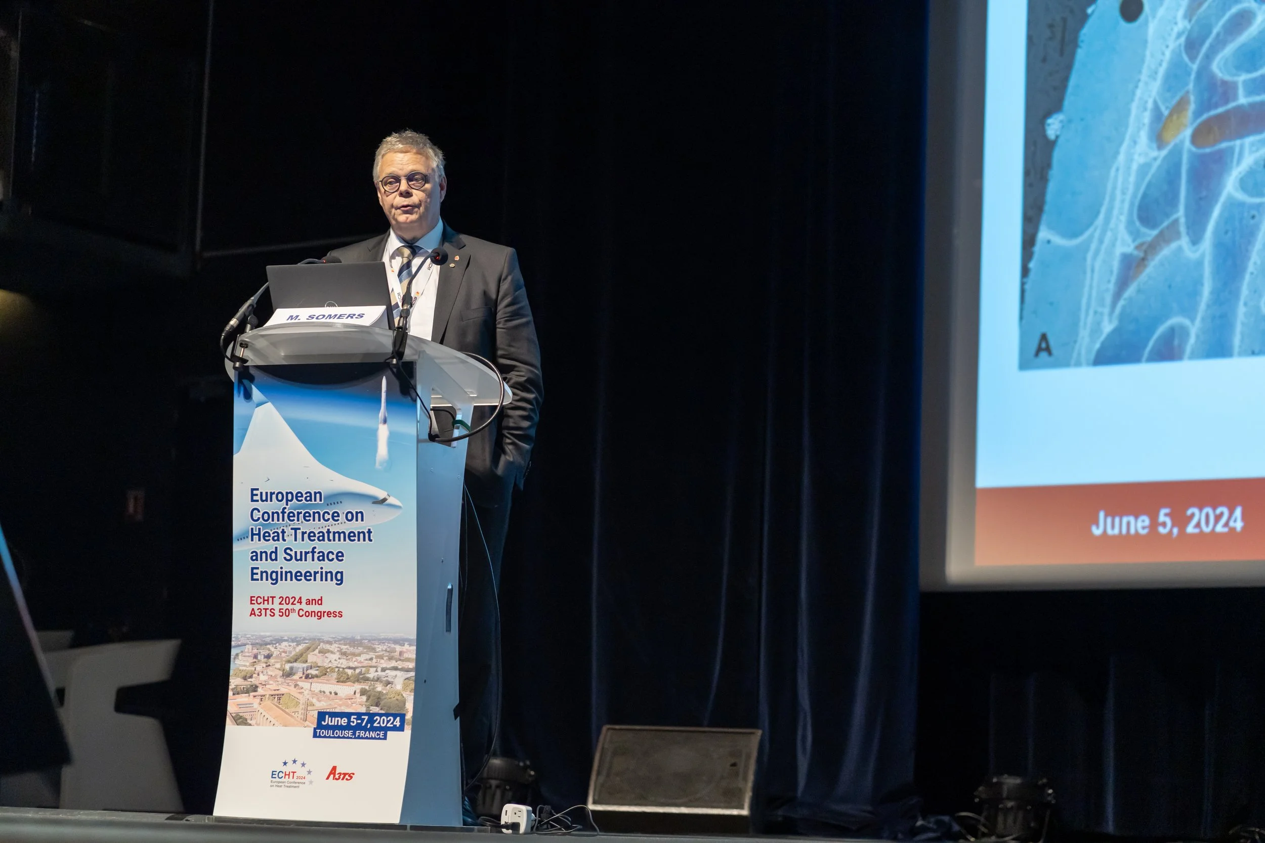 Un homme en costume présente lors de la Conférence Européenne sur le Traitement Thermique et l'Ingénierie de Surface. Un podium affiche des informations sur l'événement ayant lieu à Toulouse, France, du 5 au 7 juin 2024.