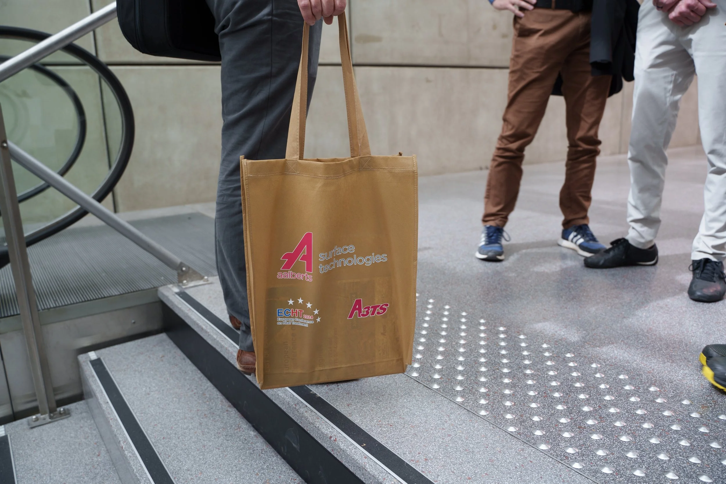 Trois personnes, vêtues de pantalons variés, se tiennent près d'un escalier. L'une d'elles tient un sac en tissu marron avec le logo "aalberts surface technologies" imprimé dessus, ainsi que d'autres logos et inscriptions. Le sol est en carrelage ave