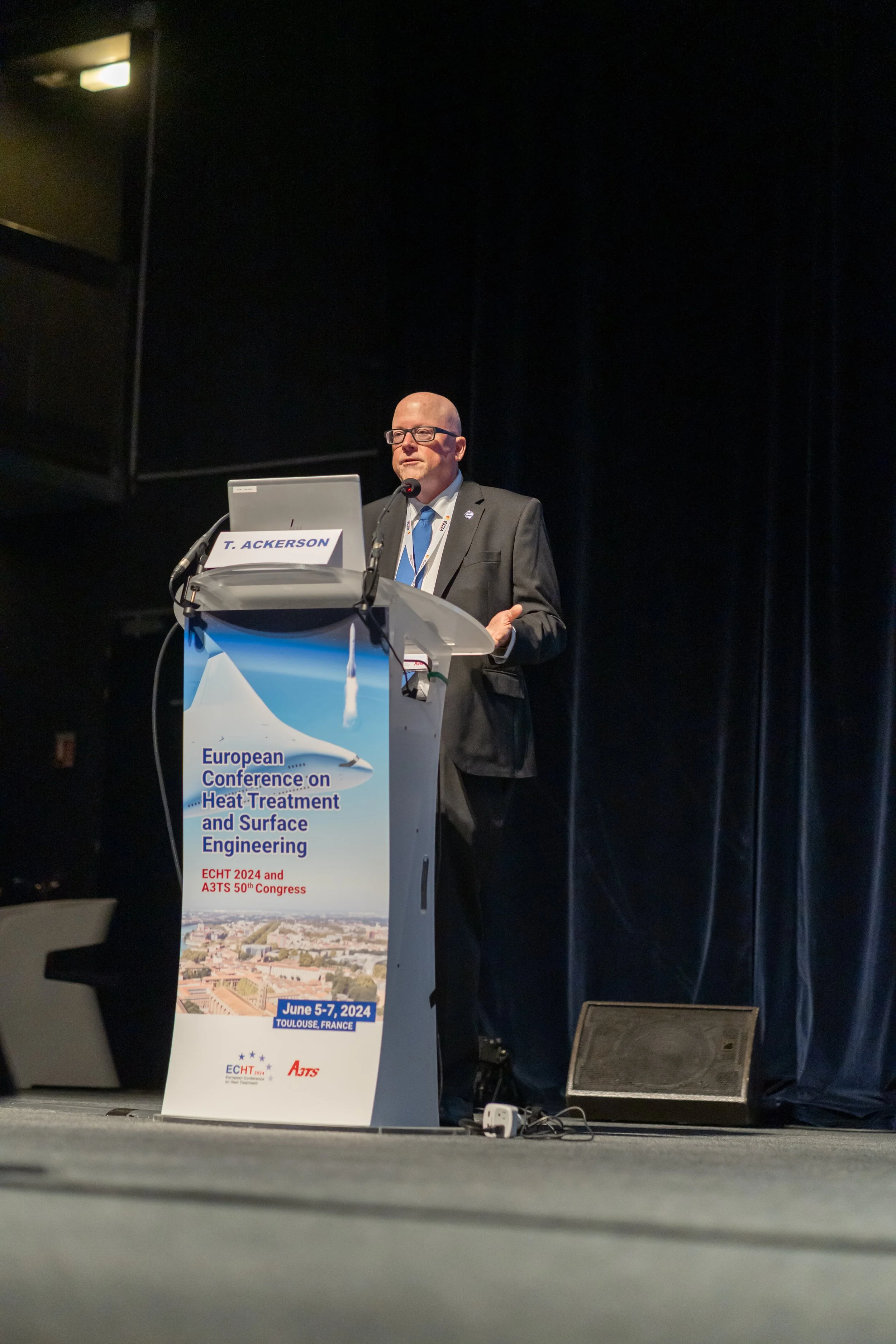 Un conférencier en costume debout derrière un pupitre lors de la conférence européenne sur le traitement thermique et l'ingénierie de surface. L'événement est programmé du 5 au 7 juin 2024 à Toulouse, France.