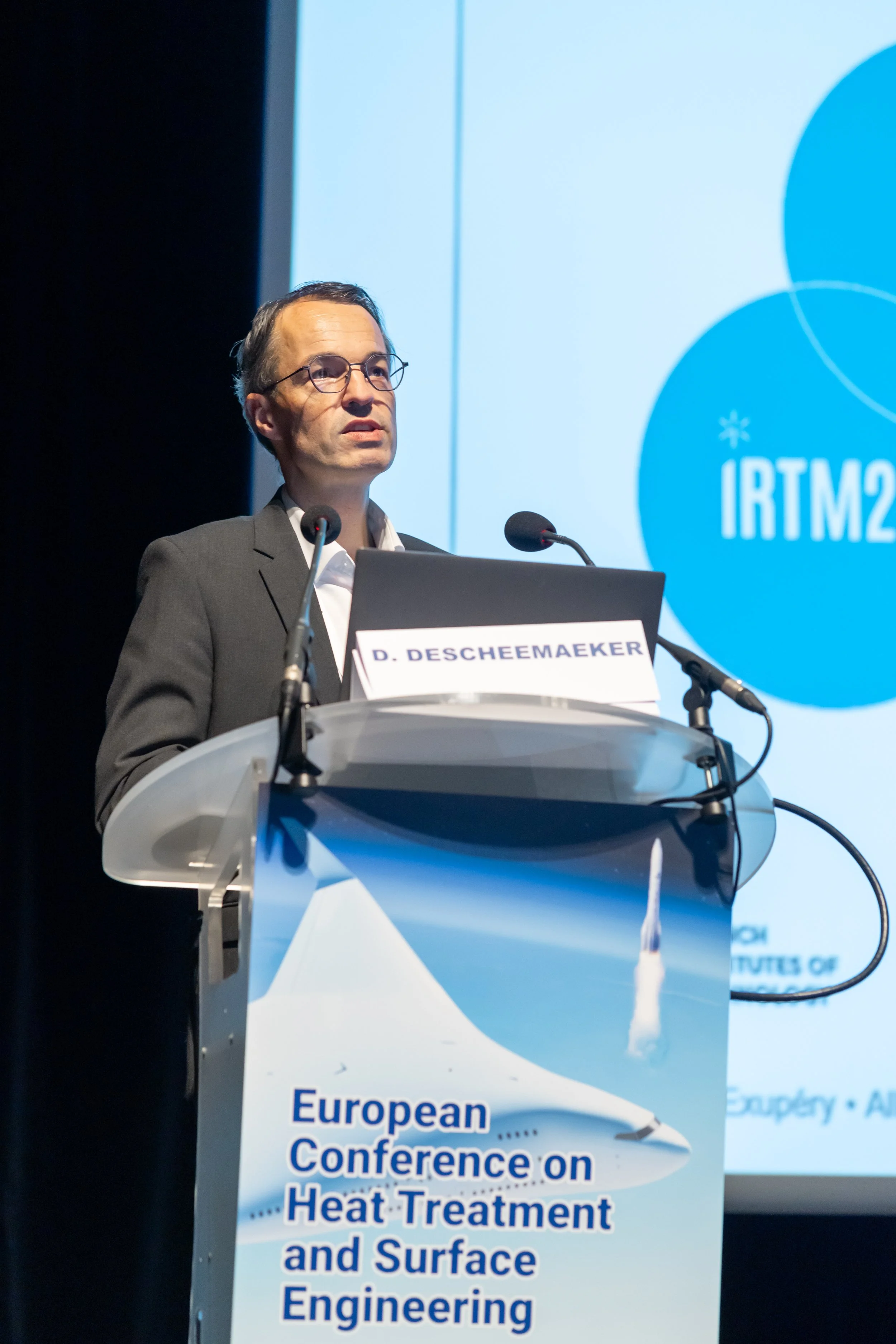 Un homme en costume parlant sur un podium lors de la Conférence Européenne sur le traitement thermique et l'ingénierie de surface, avec un panneau indiquant "IRTM2023" en arrière-plan.