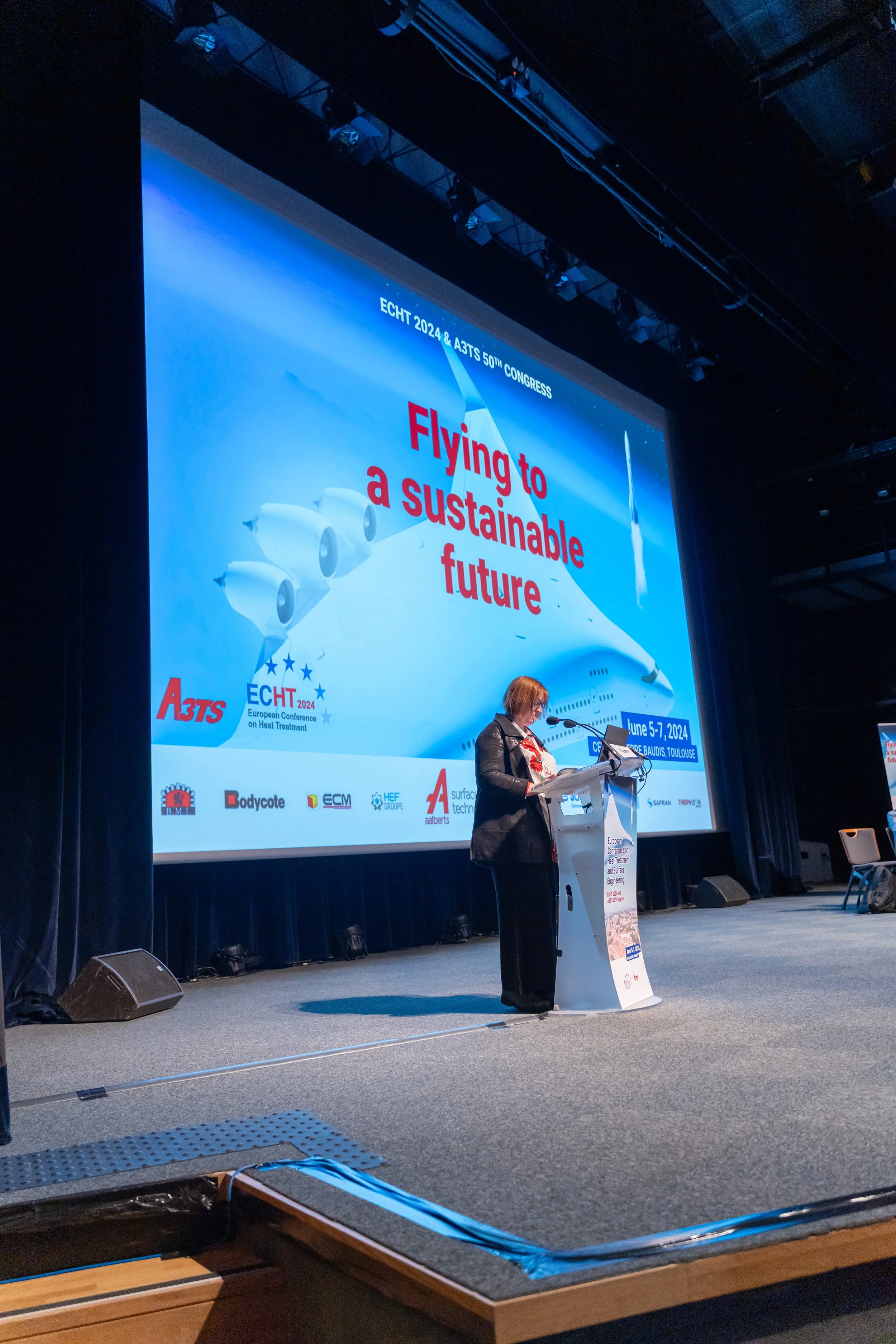 Une femme parle sur scène lors d'une conférence, avec un écran affichant "Flying to a sustainable future" et des logos d'organisations. La conférence a lieu à Toulouse du 5 au 7 juin 2024.