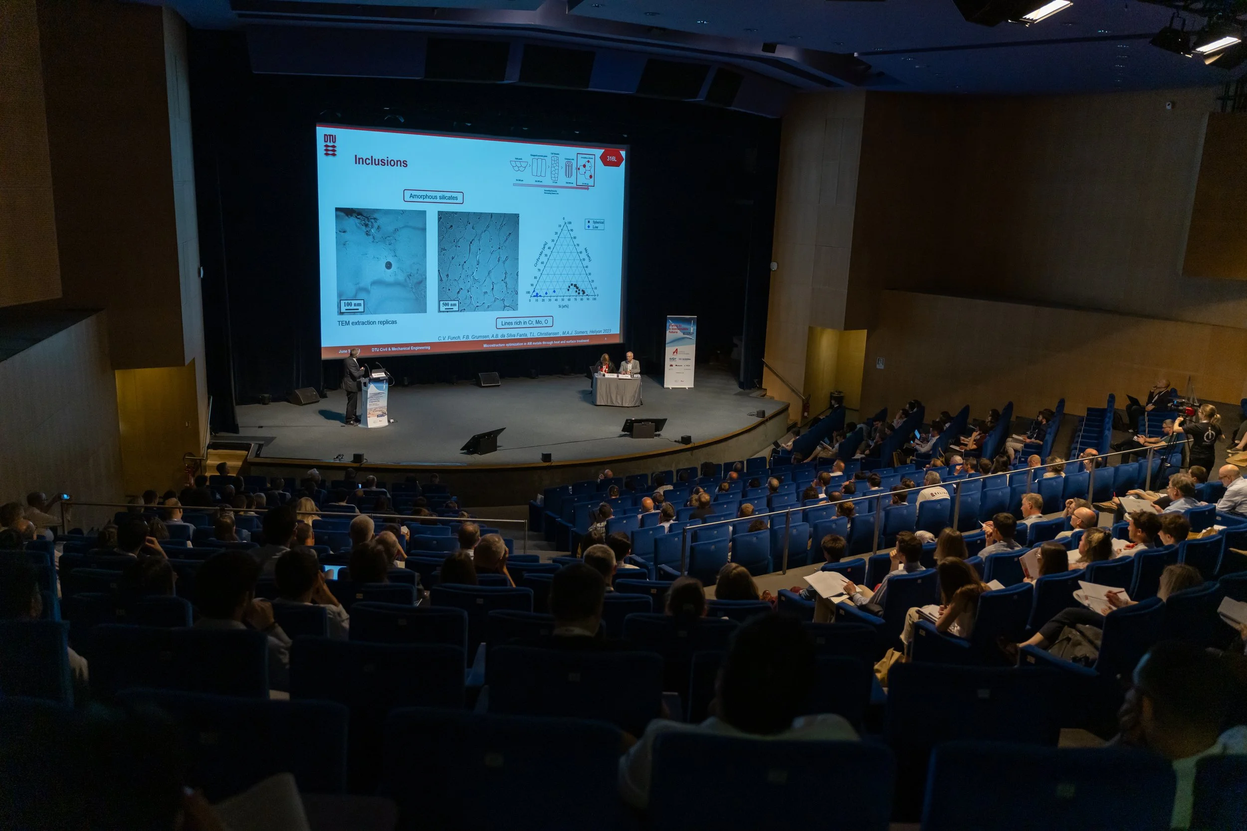 Conférence avec un orateur sur scène présentant des diapositives à un public dans un amphithéâtre.