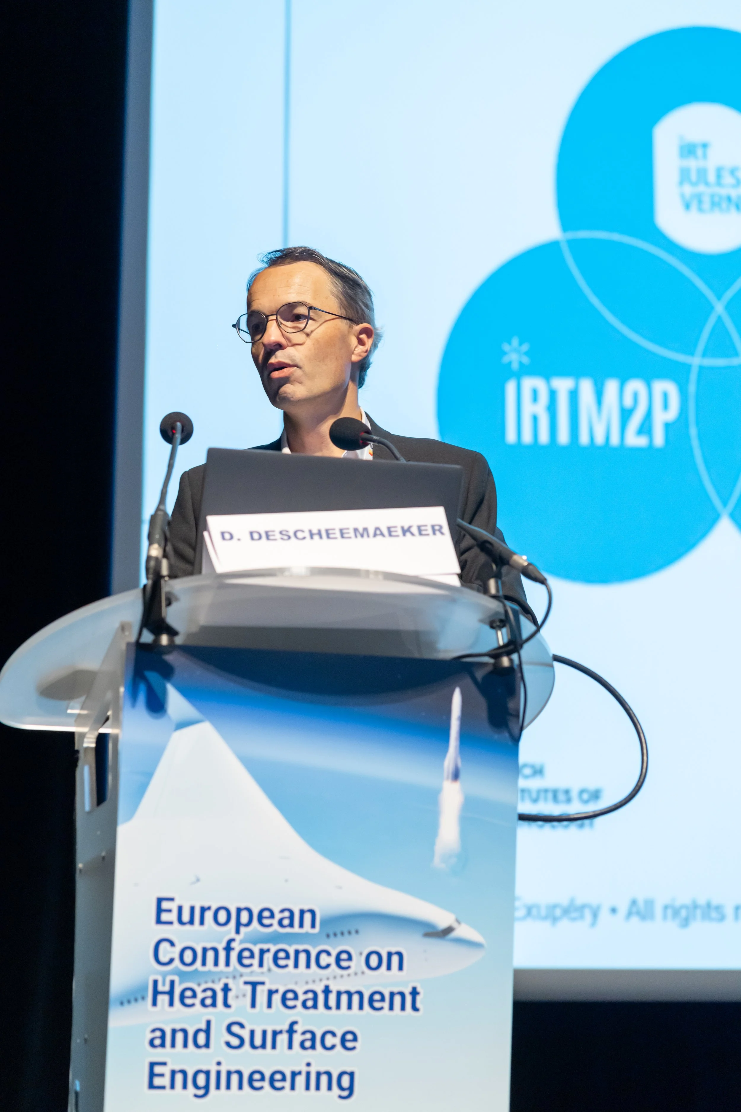 Un homme en costume parle devant un podium lors de la conférence européenne sur le traitement thermique et l'ingénierie de surface, avec un affichage de diapositives en arrière-plan.
