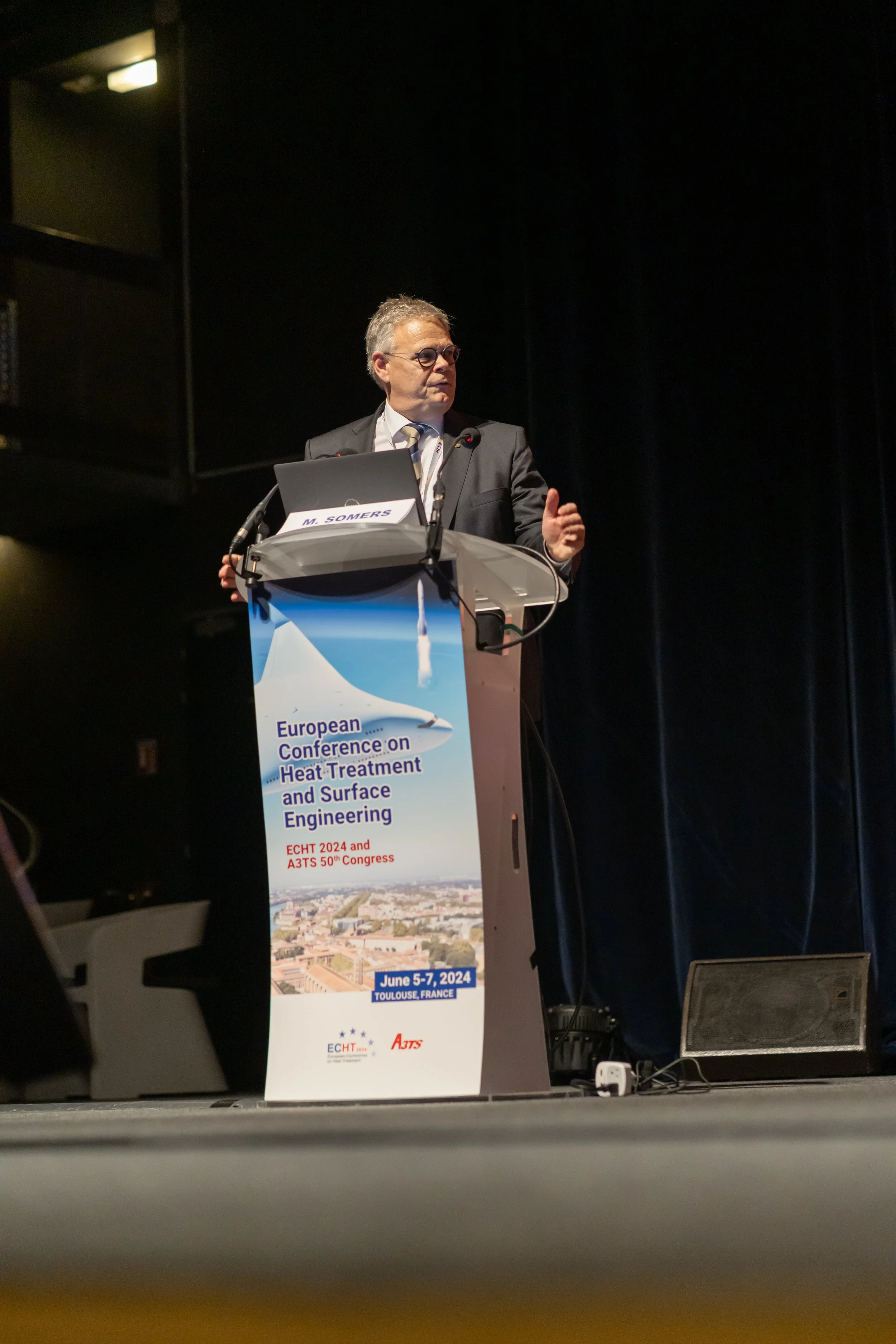Un homme en costume parle sur une scène derrière un podium avec un écran marqué "European Conference on Heat Treatment and Surface Engineering". L'événement est prévu du 5 au 7 juin 2024 à Toulouse, France.