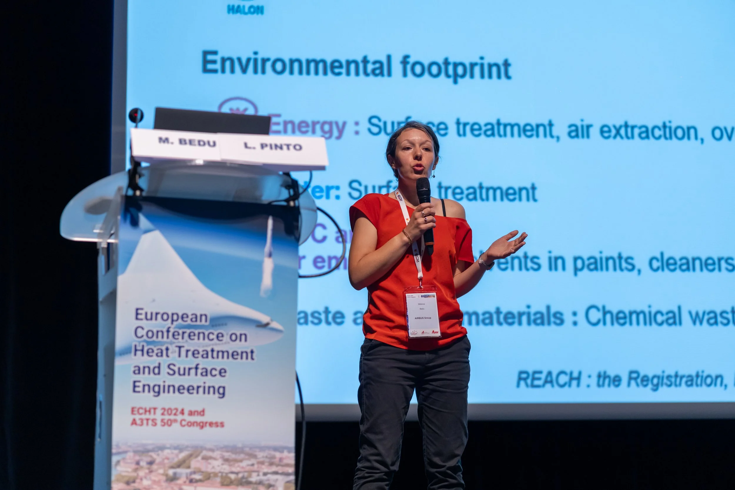 Conférencière sur scène lors d'une conférence européenne sur les traitements thermiques et le génie de surface, avec projection de diapositives sur l'empreinte écologique.