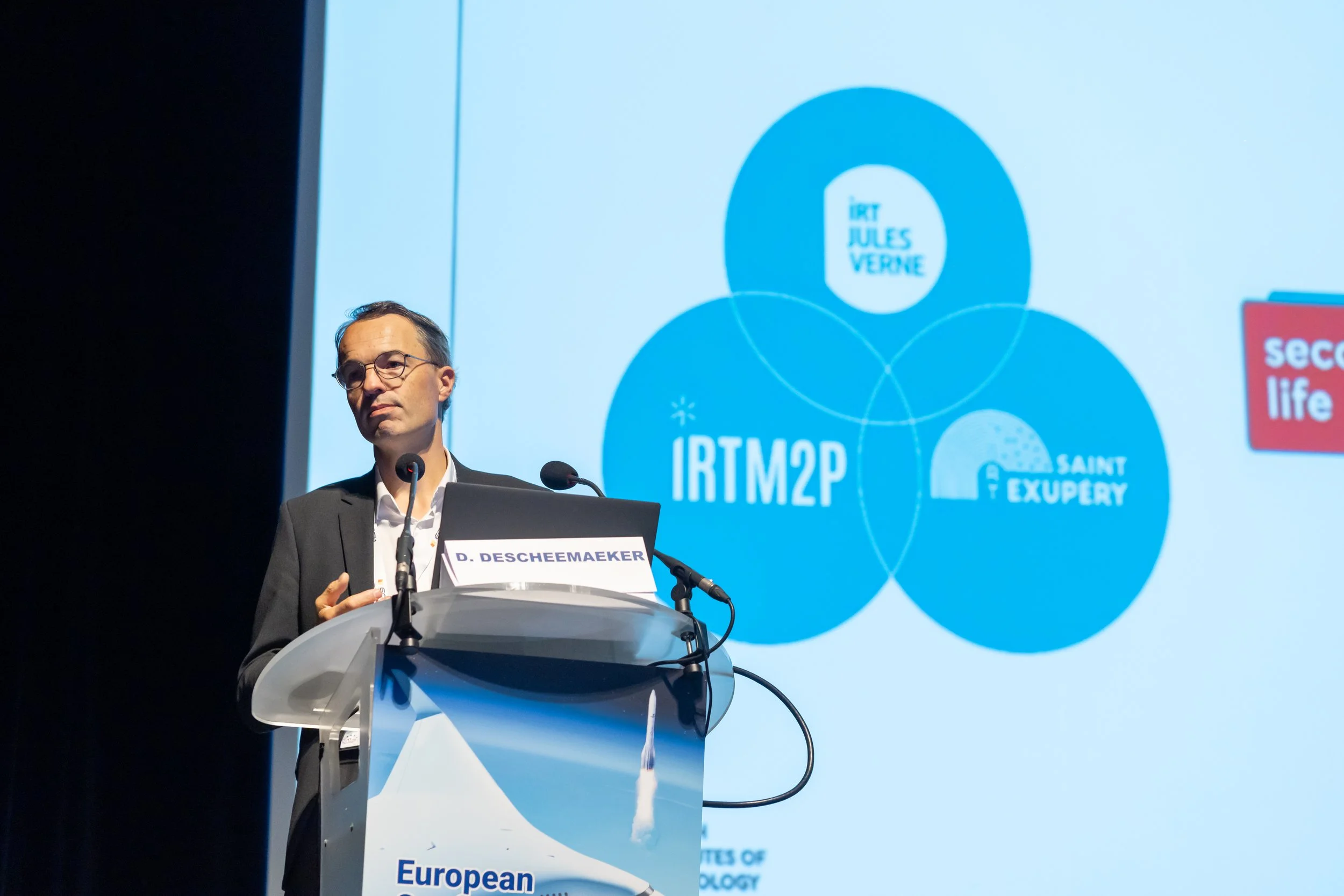 Un homme en costume présente lors d'une conférence, debout derrière un pupitre, avec un écran affichant des logos et le texte "IRTM2P" en arrière-plan.