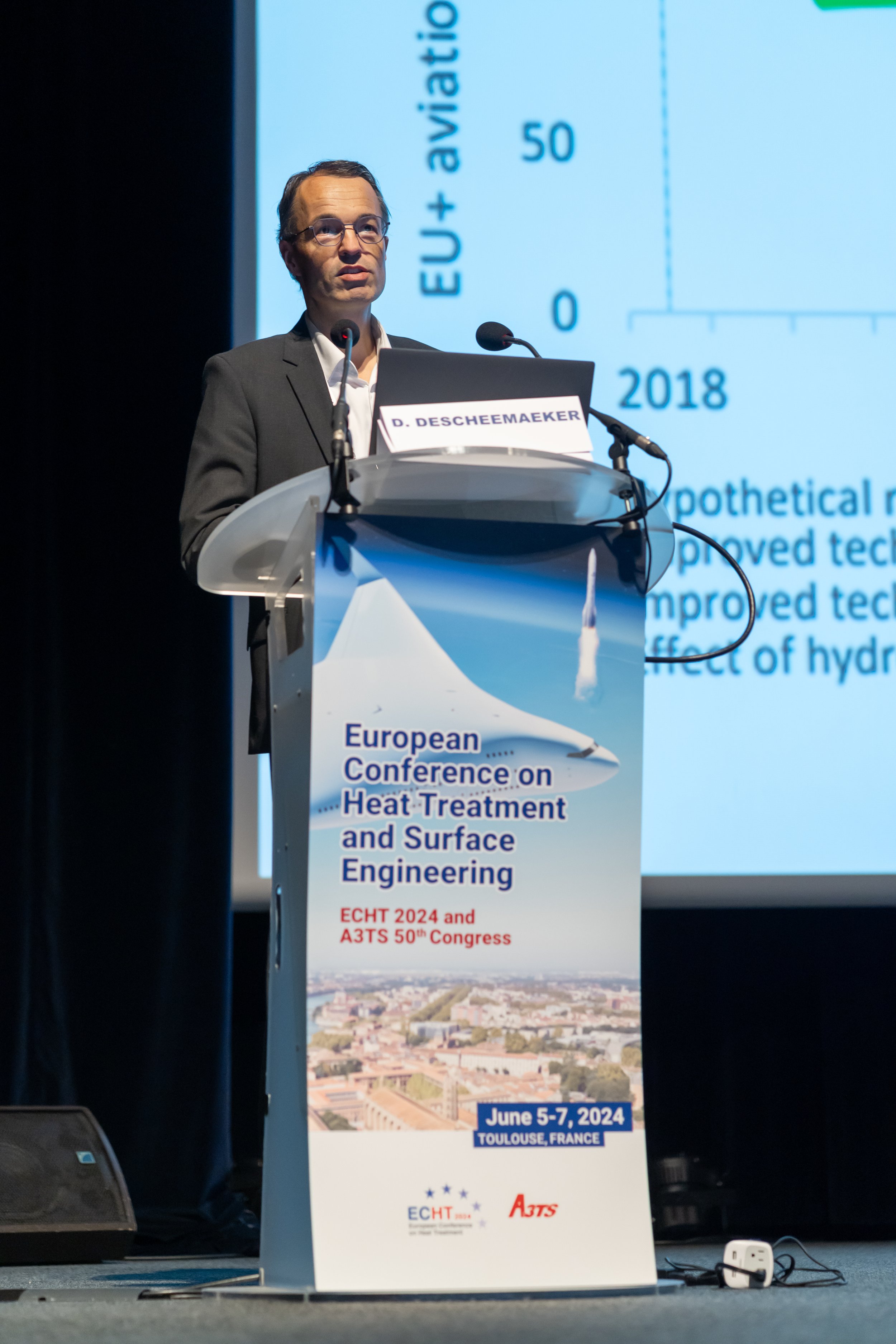 Un conférencier en costume parle sur un podium lors de la conférence européenne sur le traitement thermique et l'ingénierie de surface à Toulouse, France, du 5 au 7 juin 2024.