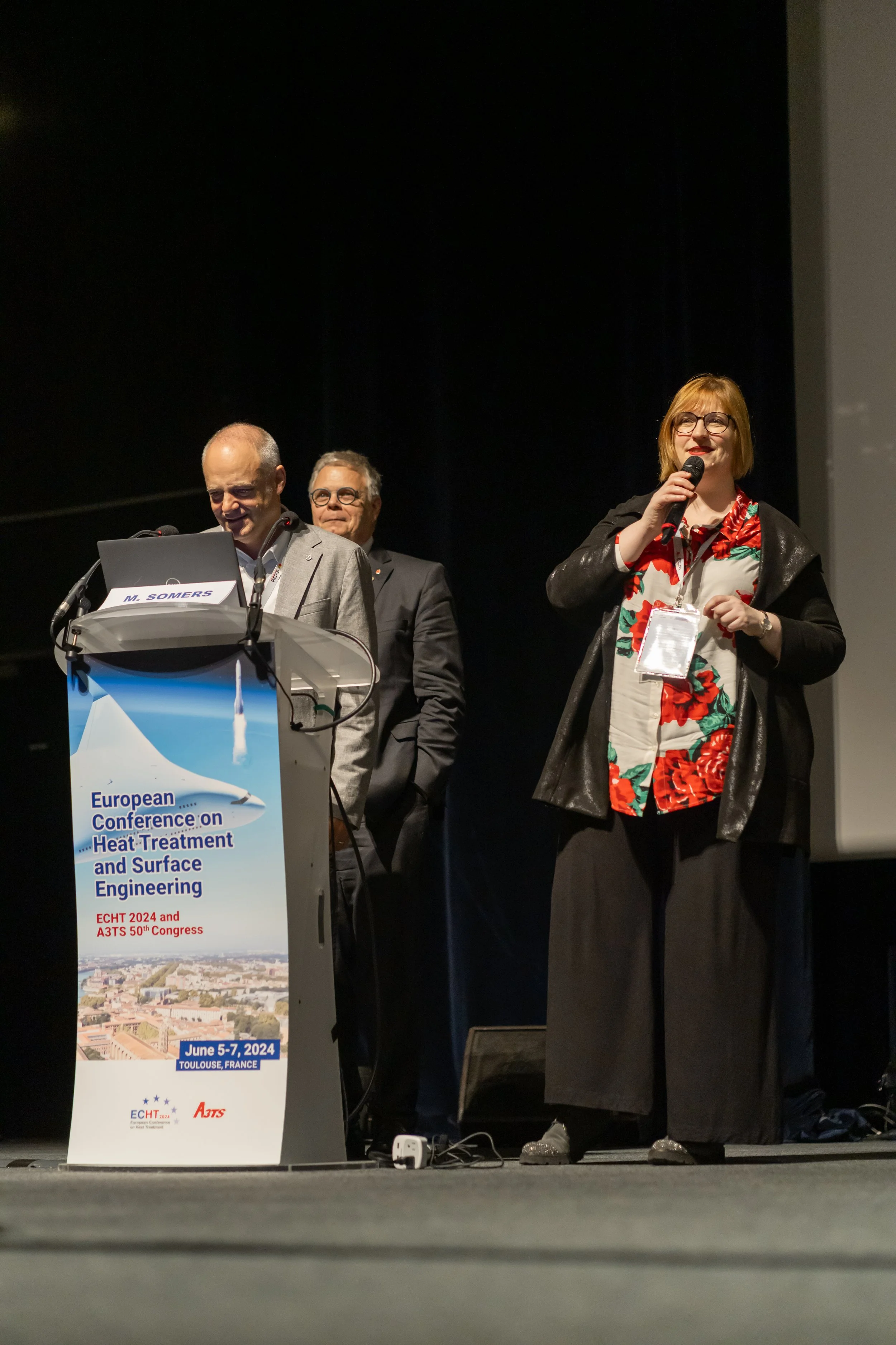 Des personnes sur scène lors d'une conférence européenne sur le traitement thermique et l'ingénierie de surface, affichant un grand panneau publicitaire pour l'événement prévu en juin 2024 à Toulouse, France.