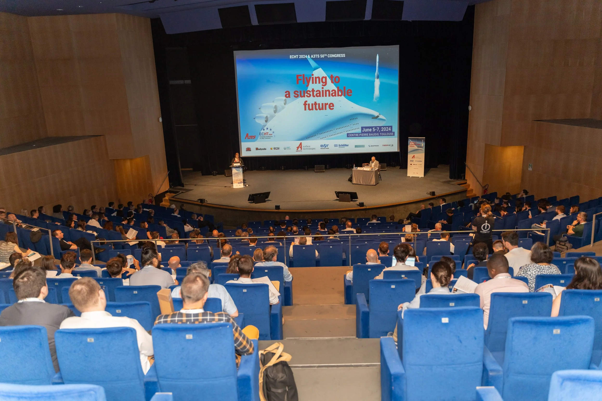 Une conférence dans un amphithéâtre avec une présentation intitulée "Flying to a sustainable future" affichée sur grand écran. De nombreuses personnes sont assises dans des fauteuils bleus, écoutant le présentateur.