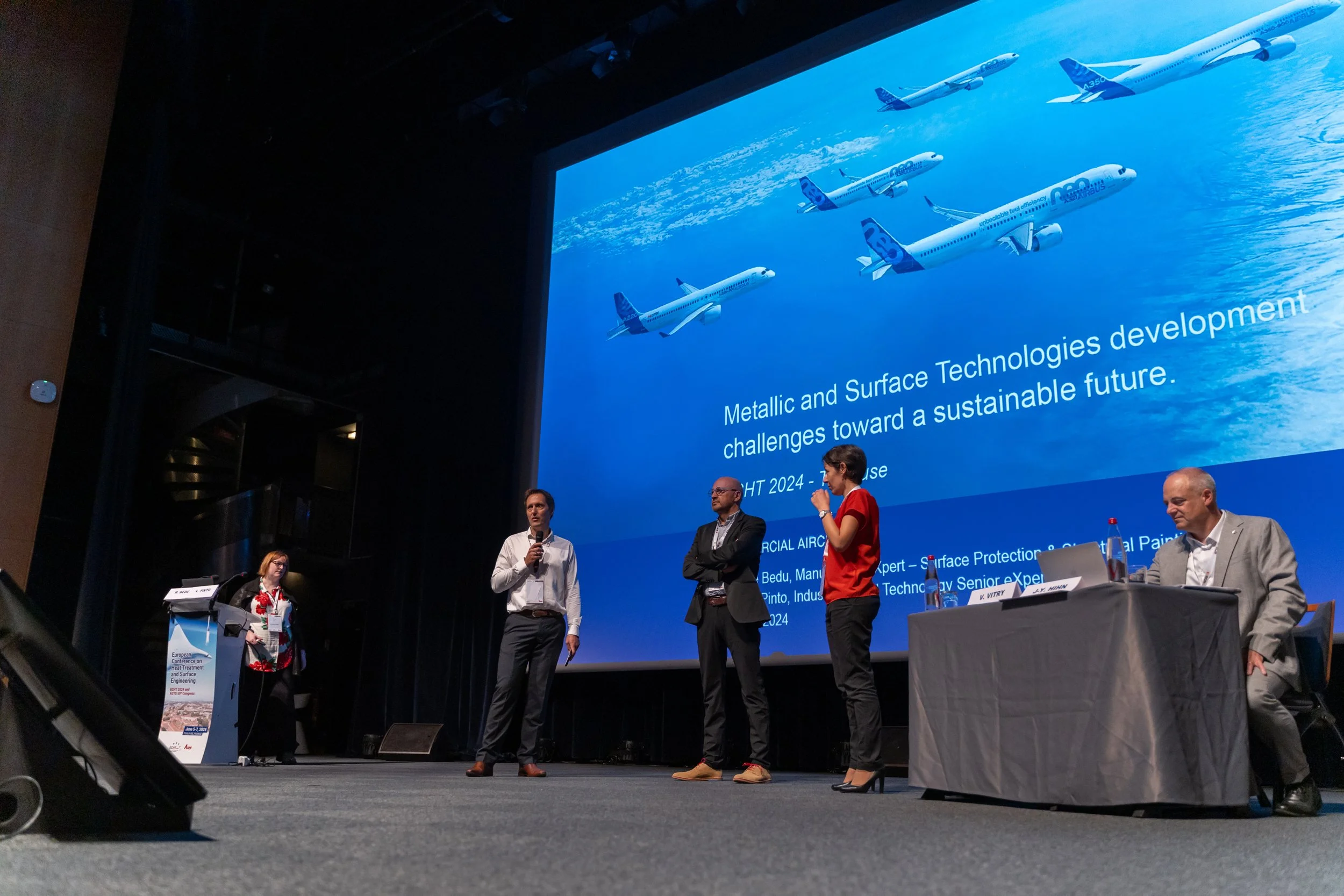 Conférence avec des intervenants sur scène et un écran affichant le texte "Développement des technologies métalliques et de surface, défis vers un avenir durable", avec une image d'avions en vol.