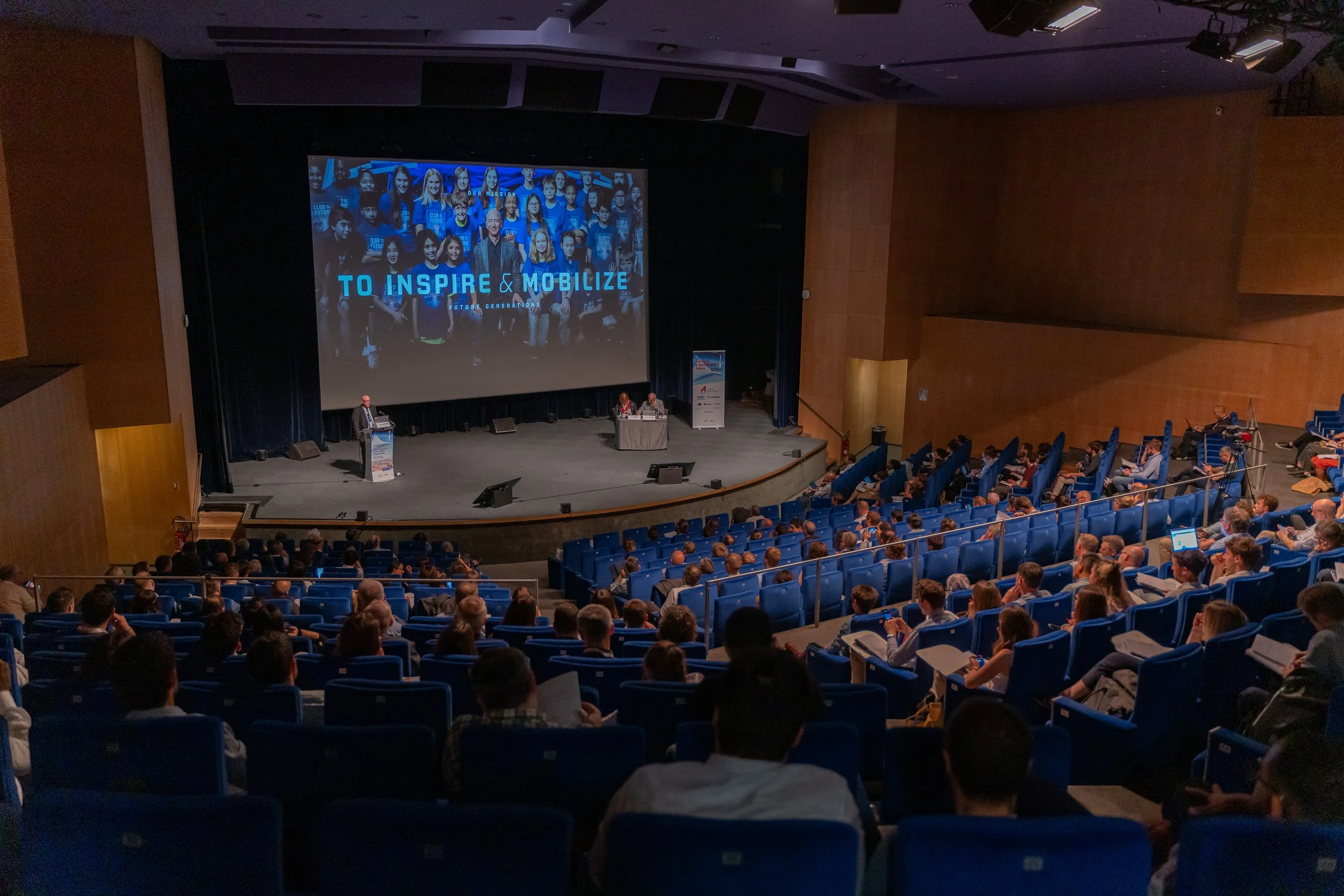 Une conférence se déroule dans un auditorium, avec un orateur s'exprimant sur scène et un grand écran affichant "To Inspire & Mobilize" devant un public assis. Les sièges sont bleus et la salle est bien remplie.