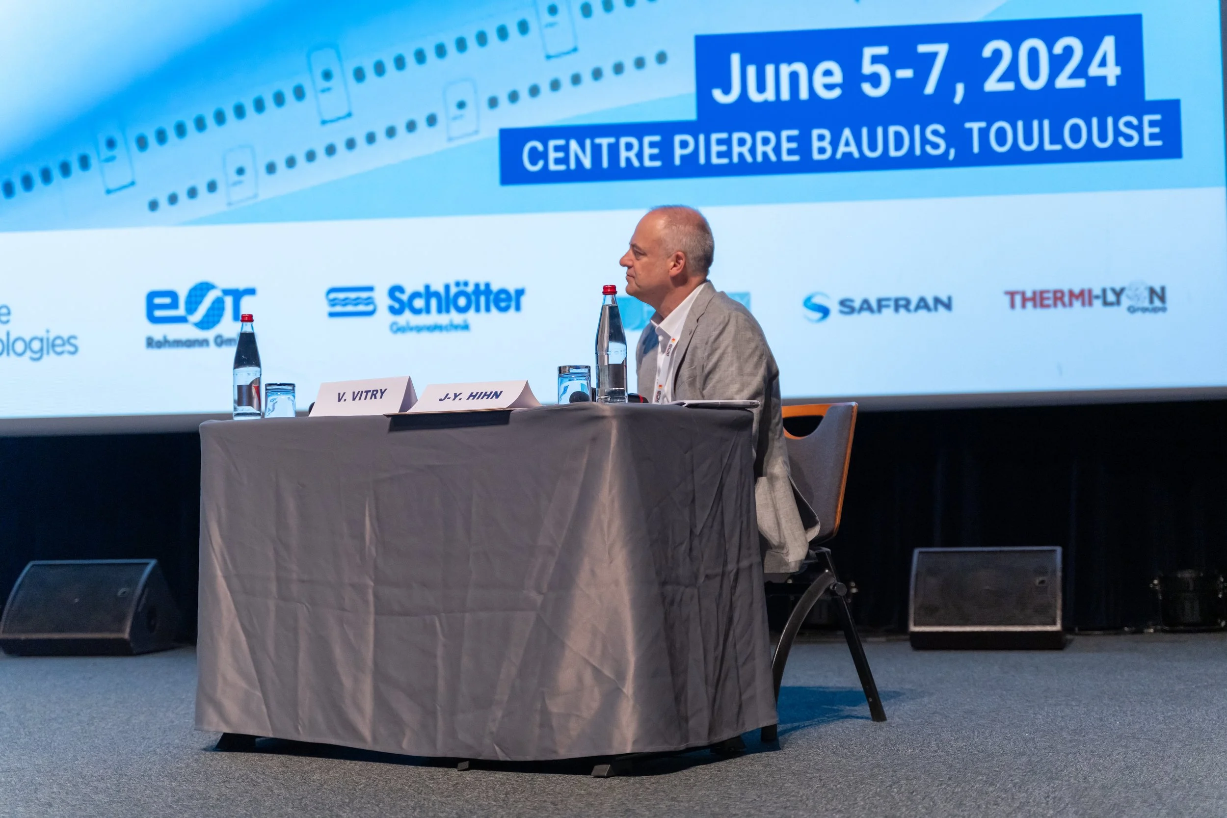 Un homme assis à une table de conférence avec un écran affichant "June 5-7, 2024, Centre Pierre Baudis, Toulouse" et des logos d'entreprises en arrière-plan.