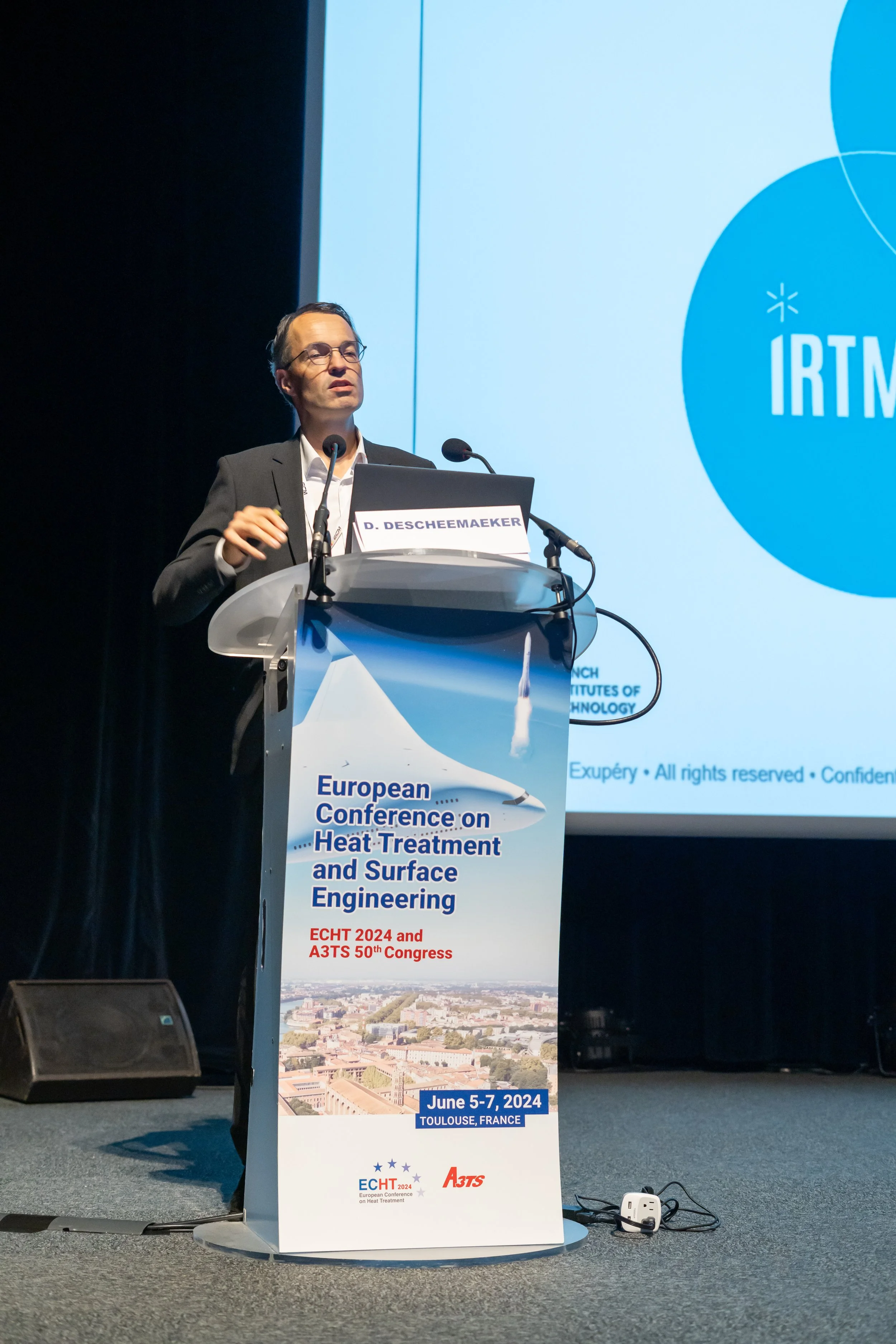 Conférencier à une conférence européenne sur le traitement thermique et l'ingénierie de surface, affichant un panneau pour l'événement prévu en juin 2024 à Toulouse, France.